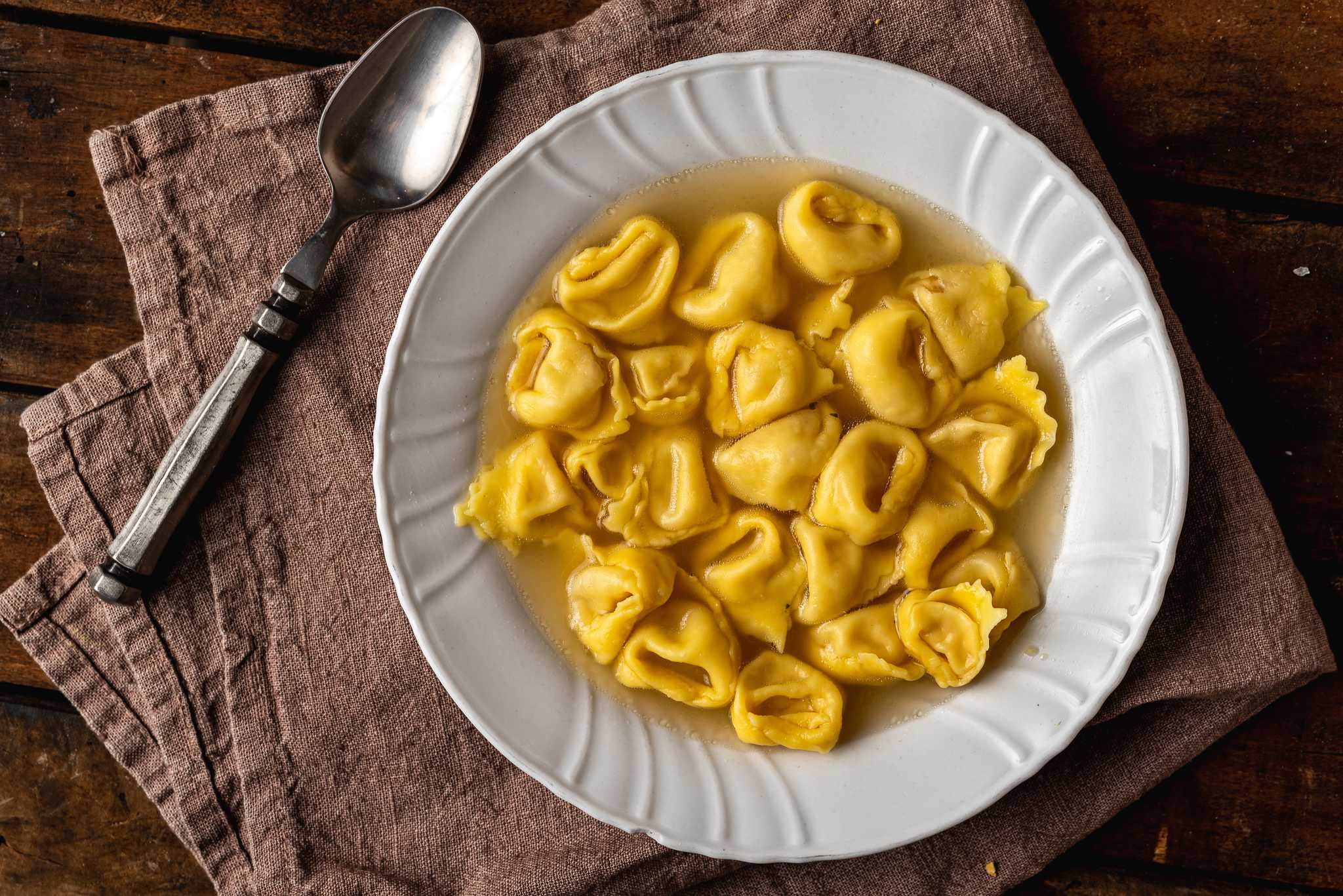 White bowl of tortellini in broth sitting on a brown cloth napkin beside a silver spoon.