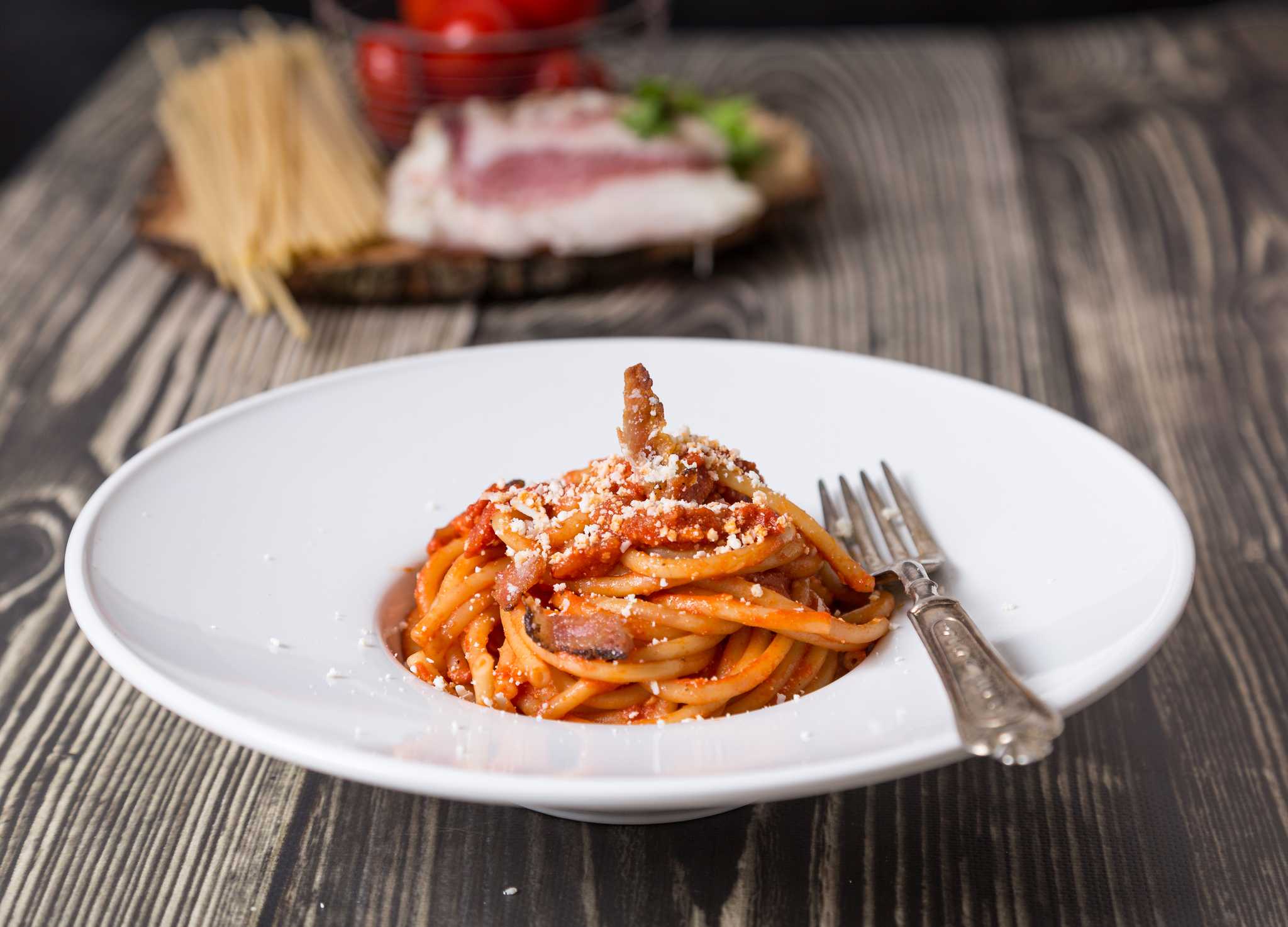 White bowl of bucatini all’Amatriciana topped with grated cheese, a silver fork resting on the rim.
