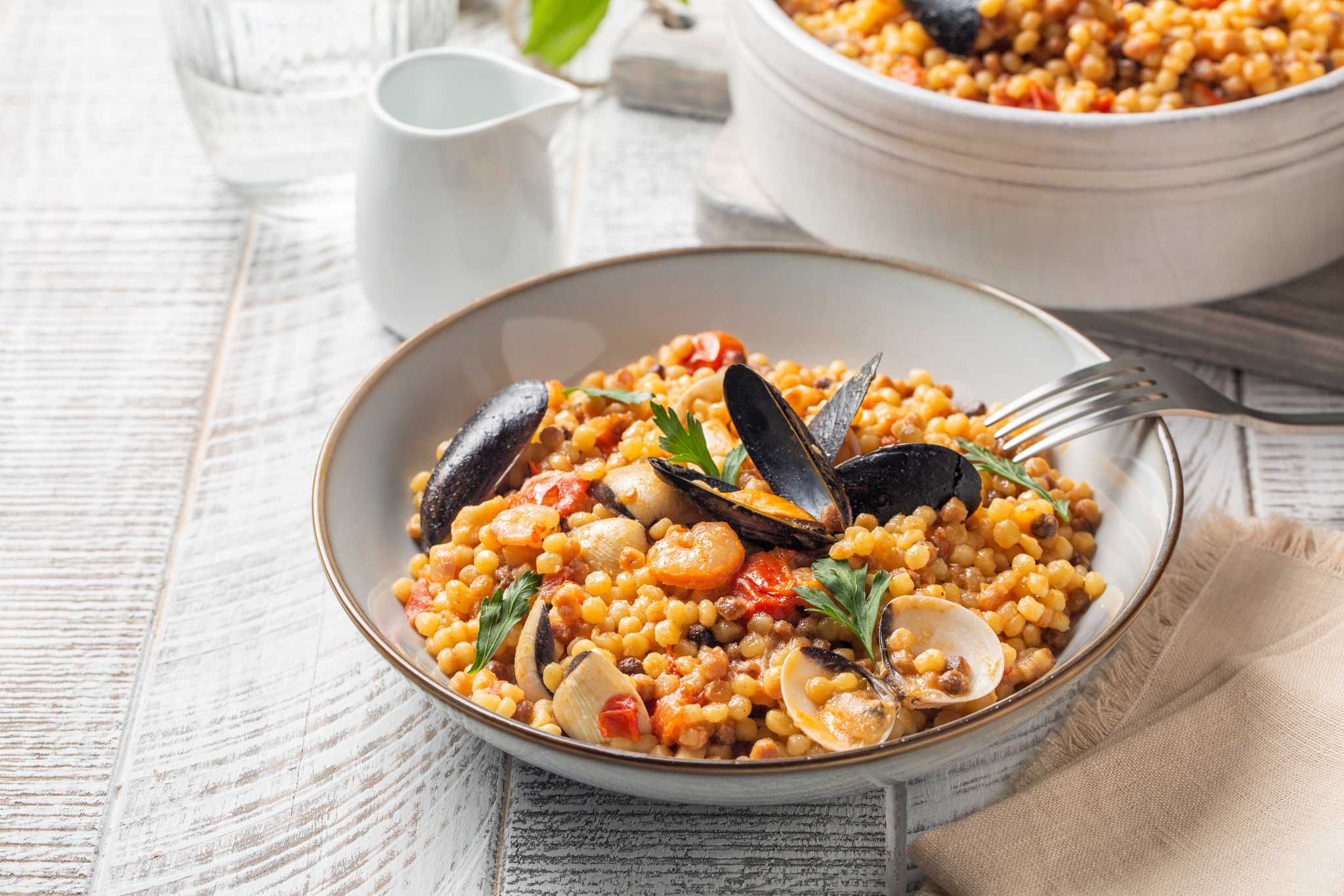 White bowl of fregola with mussels and other seafood, a fork propped on the bowl.
