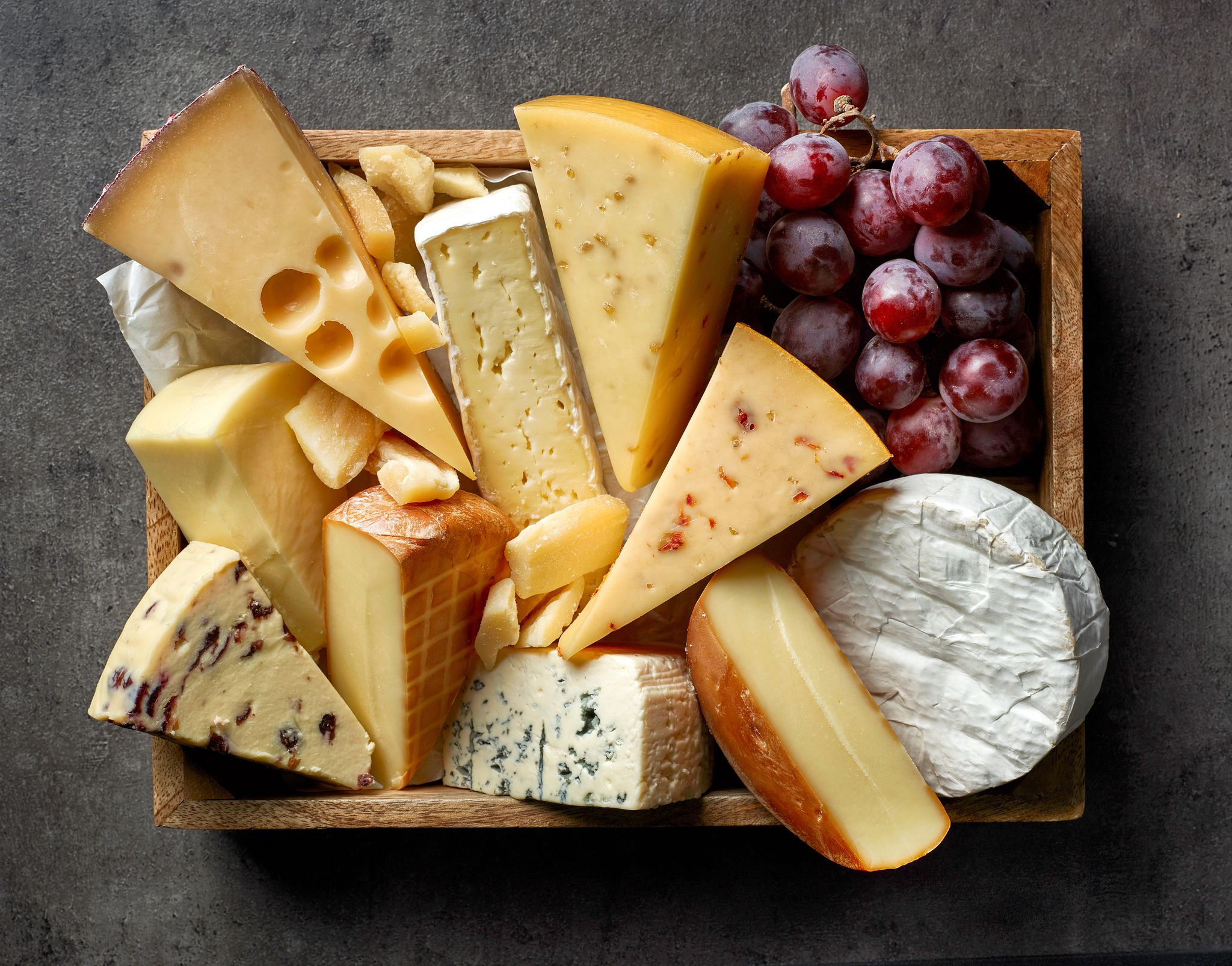 Rectangular wooden platter of various wedges of cheese and red grapes.