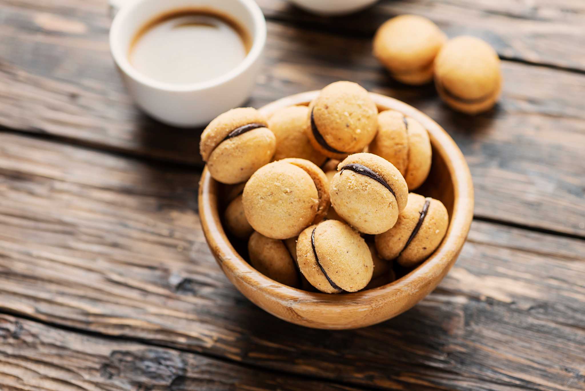 Wooden bowl of baci di dama beside a cup of coffee.