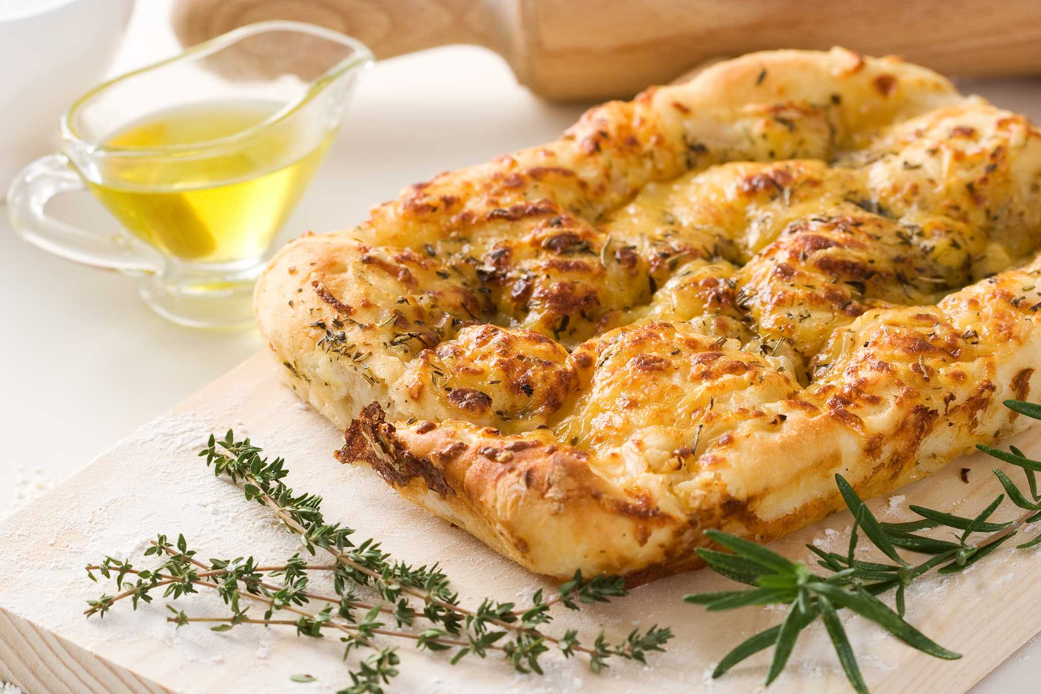 Focaccia topped with herbs, beside sprigs of herbs and a glass container of oil.