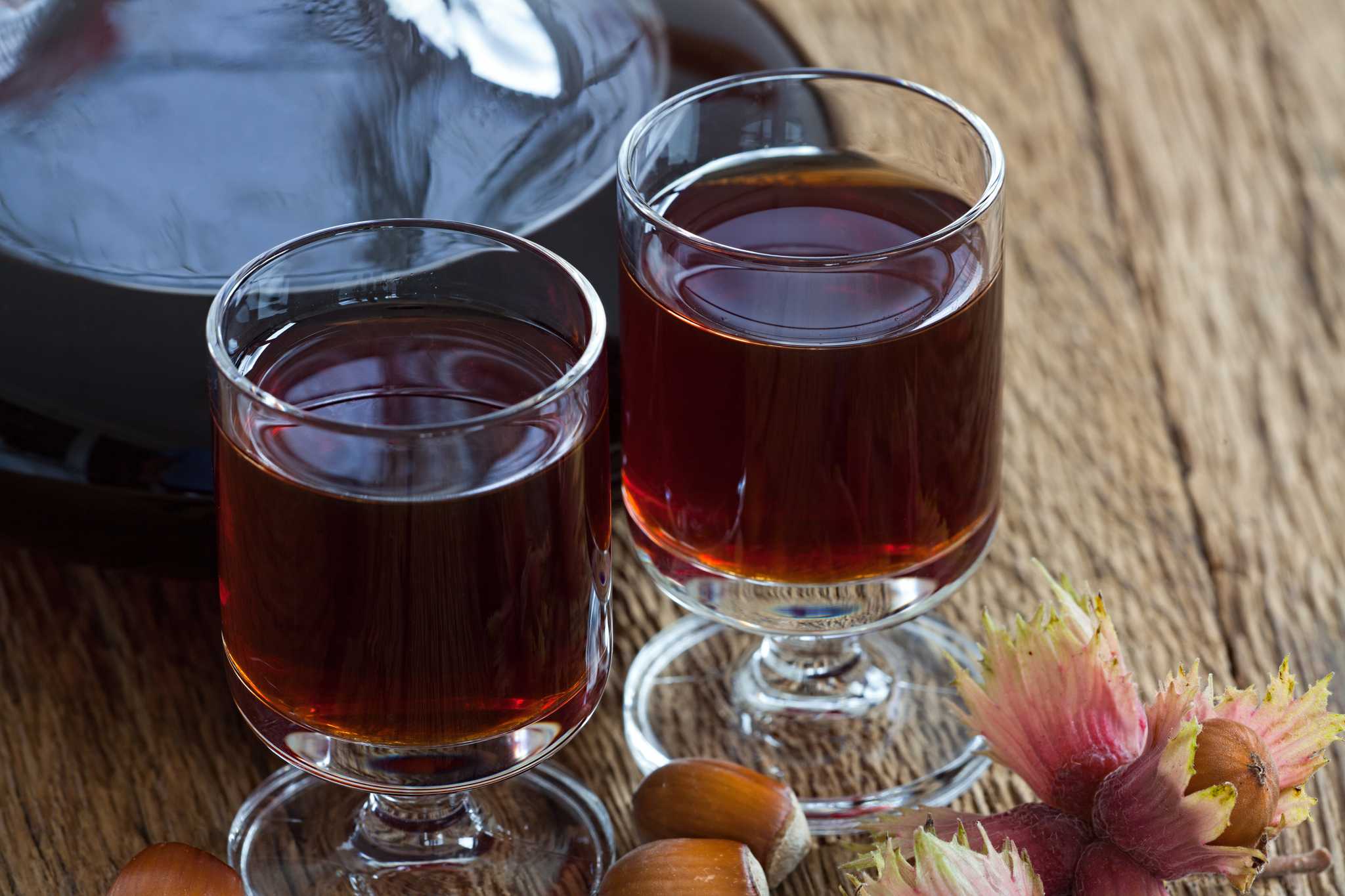 Two small glasses of brown liquor with nuts and flowers in the foreground.