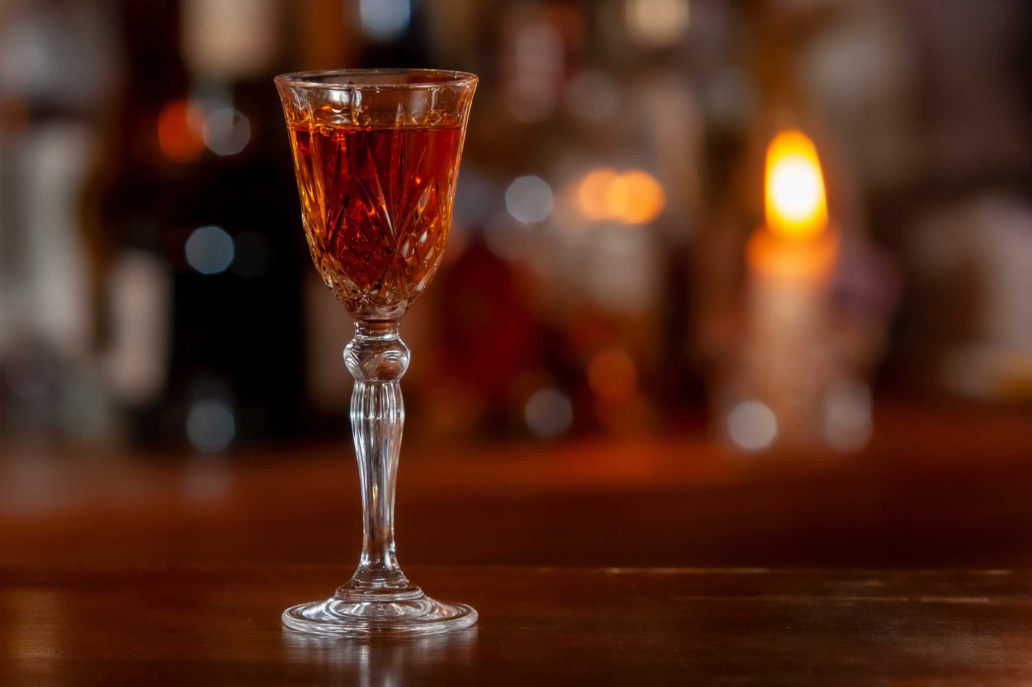 Crystal glass with long stem filled with red-brown liqueur sitting on a bar.