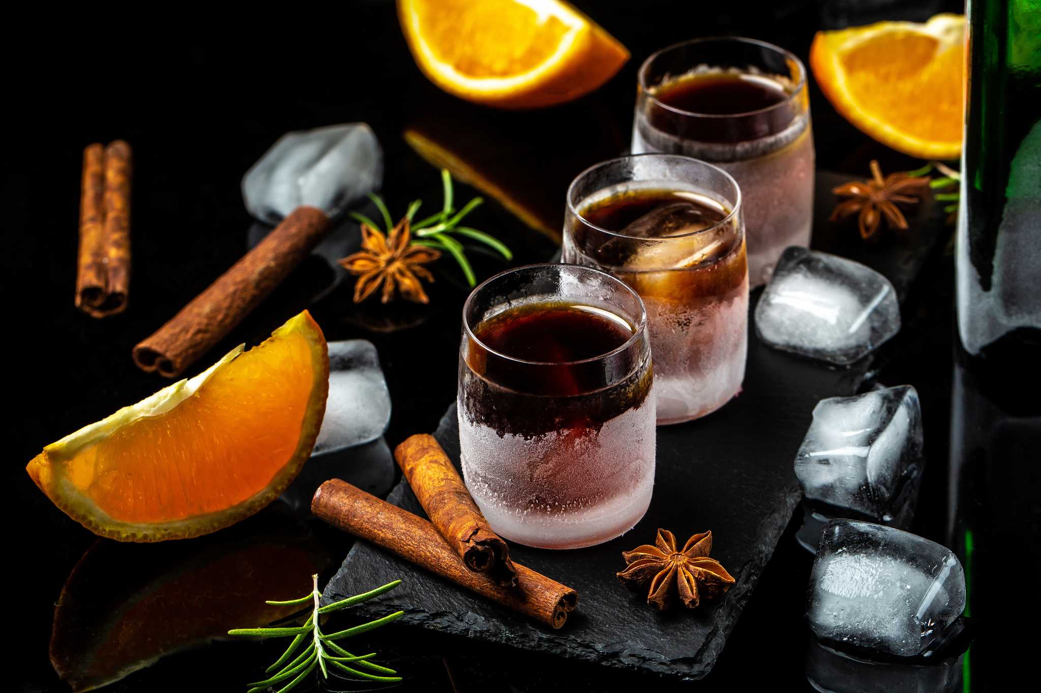Three small glasses of sambuca surrounded by ice, orange slices, star anise, and cinnamon sticks.