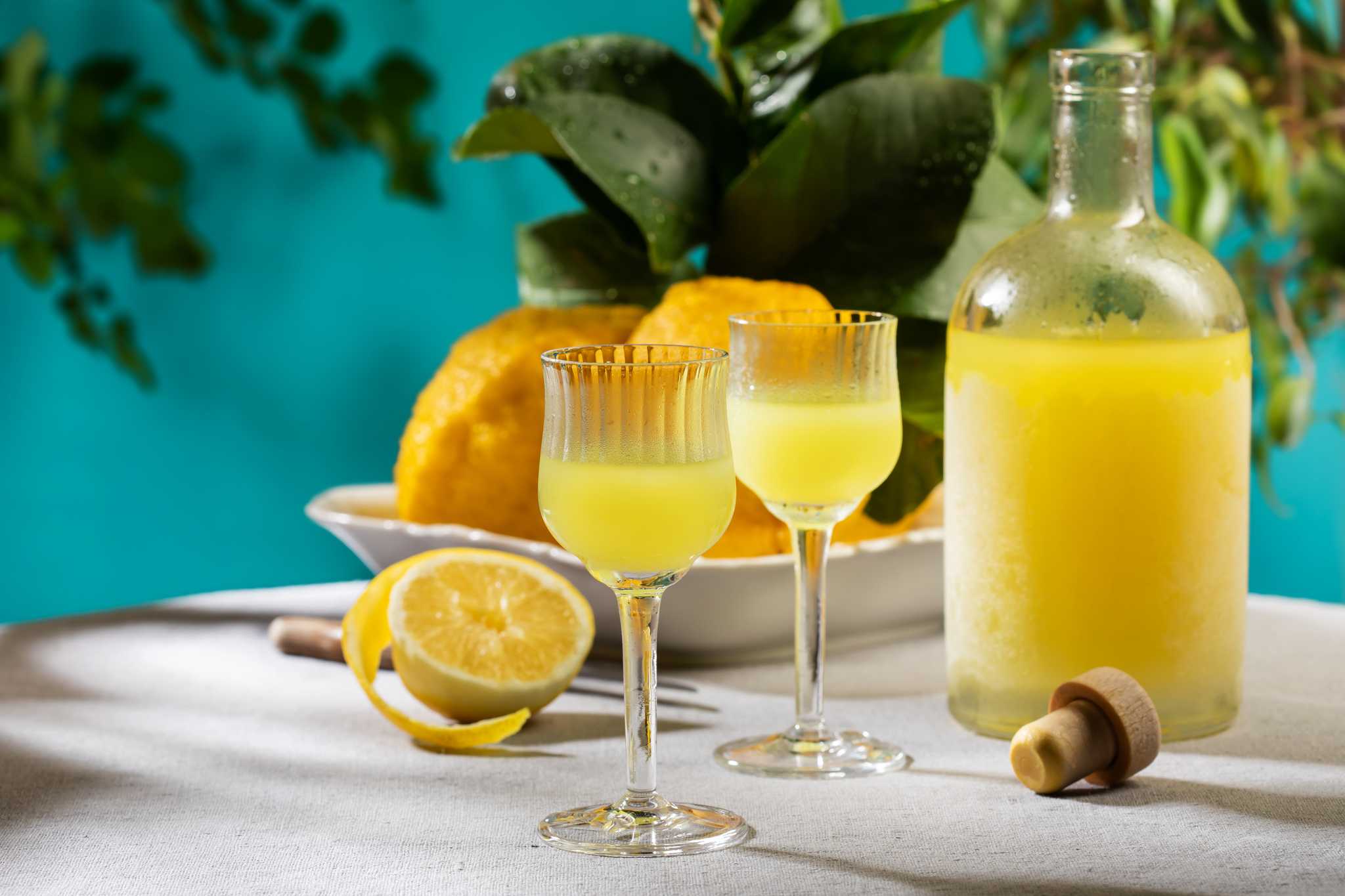 Two glasses and an uncorked bottle of limoncello on a table, with a bowl of lemons behind.