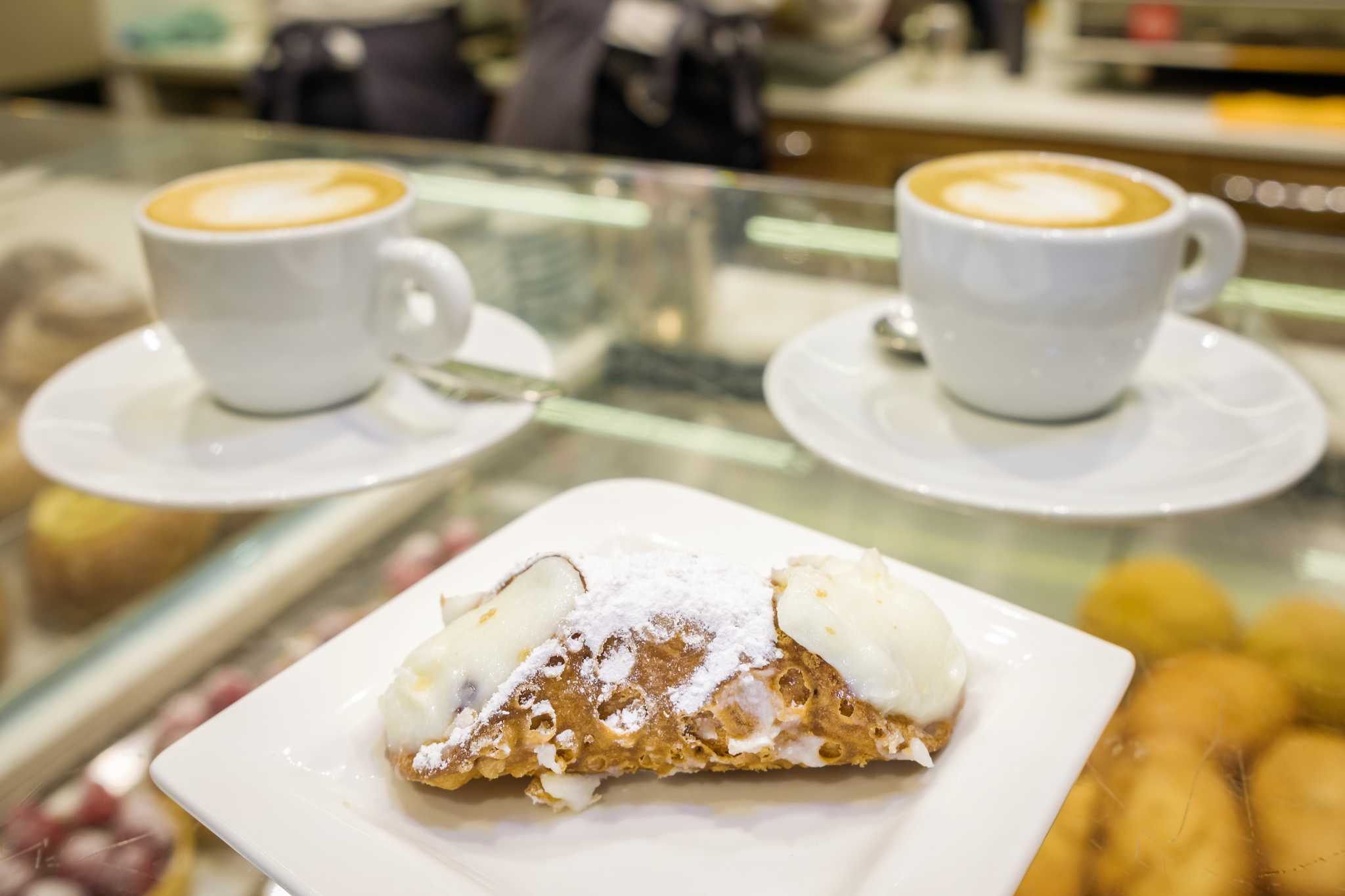 Two coffees in white mugs and a cannoli at a glass bar counter in Sicily.