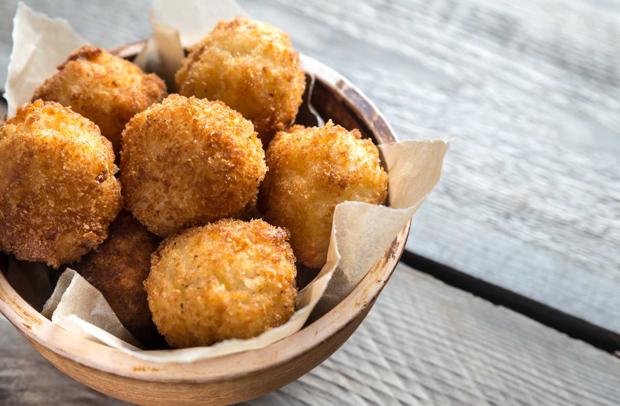 Bowl of several golden-brown arancini on a grey wooden table.