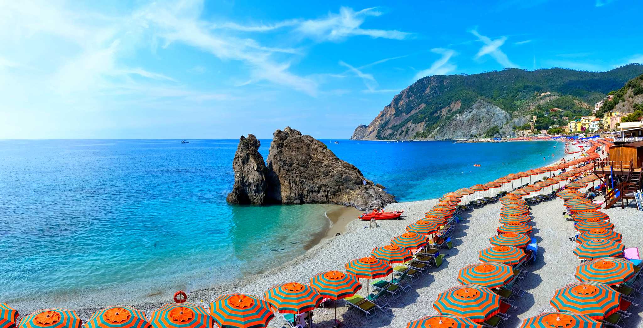 Three rows of striped umbrellas on the sand in front of the sea with mountains in the distance.