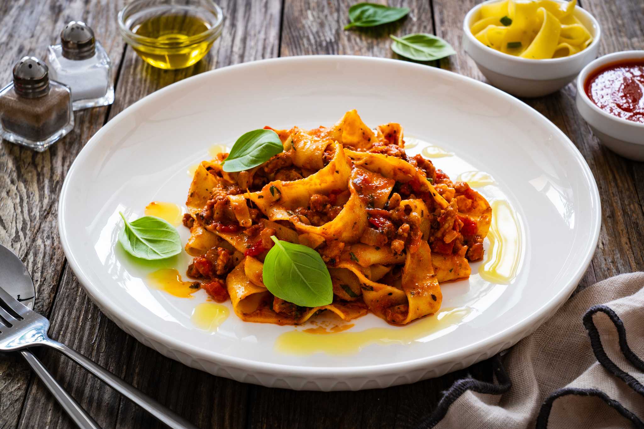Ragu alla Bolognese on a white plate on top of a wooden table, surrounded by utensils and ingredients.