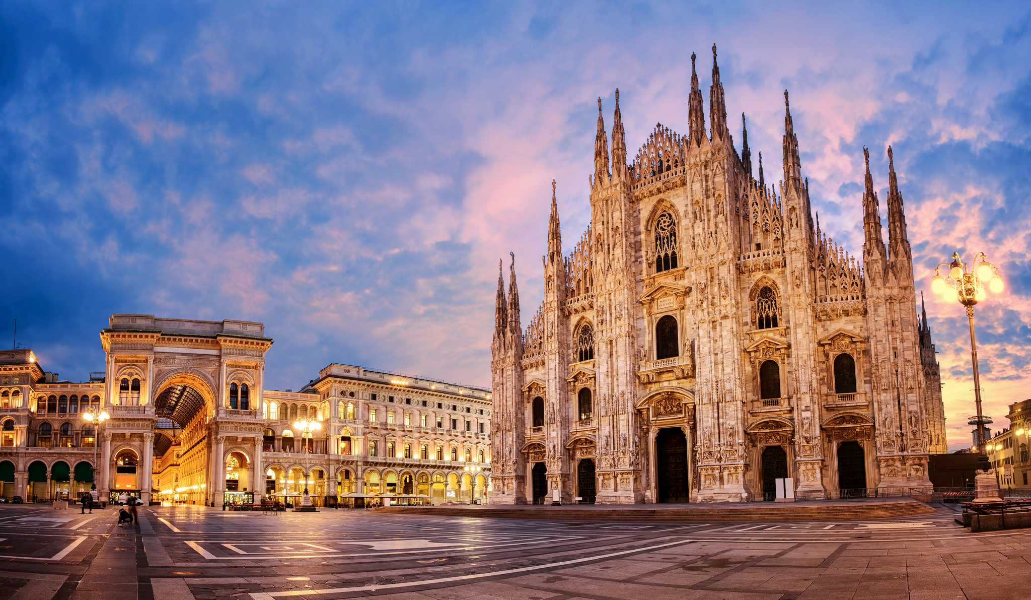 Angled view of the Milan Cathedral under a pink and blue sunset with streetlights on.