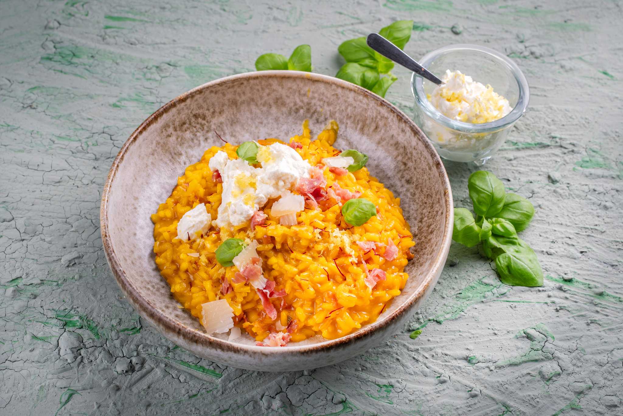 Risotto alla Milanese in a bowl with extra cheese and basil leaves on the side.
