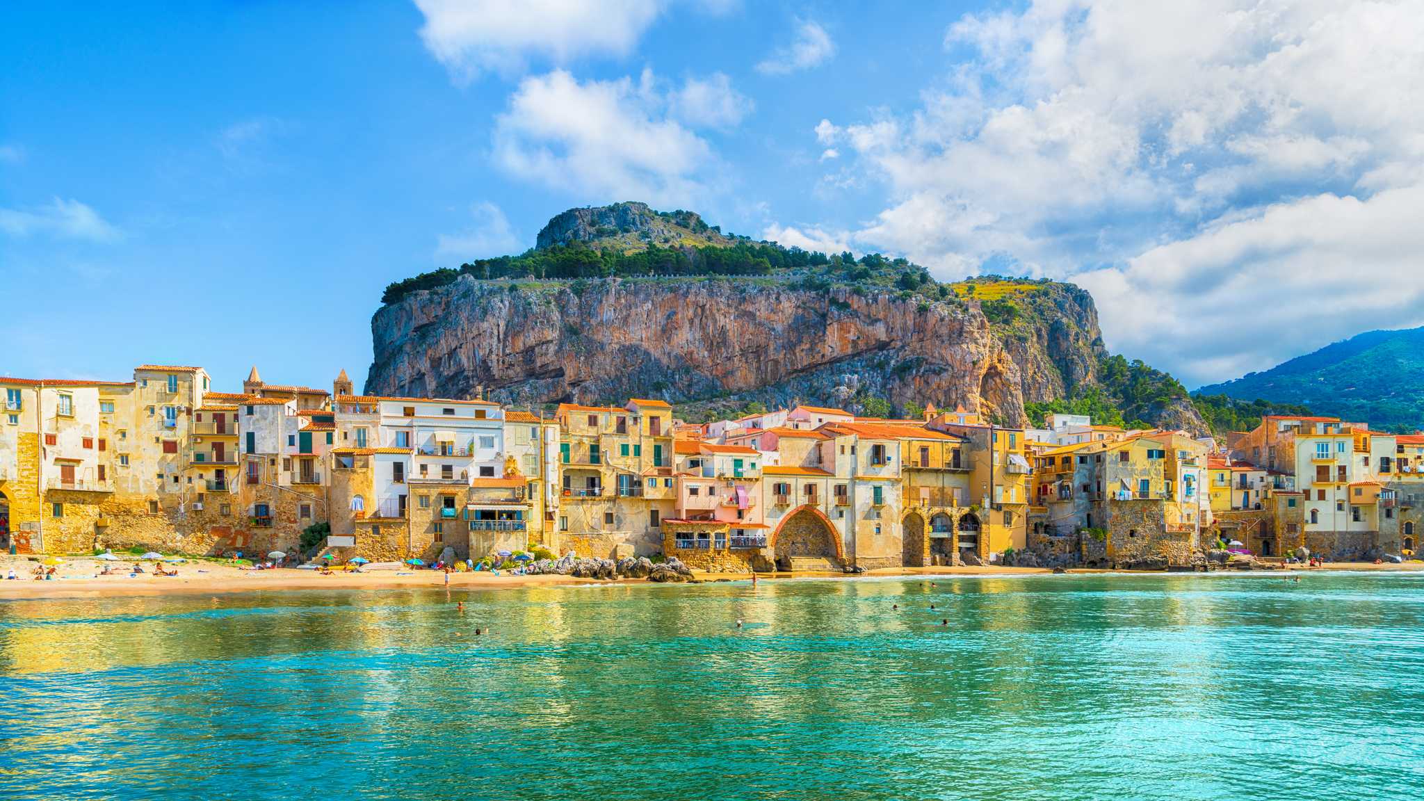 Bright blue water leading to a Sicily beach backed by buildings and a mountain face.