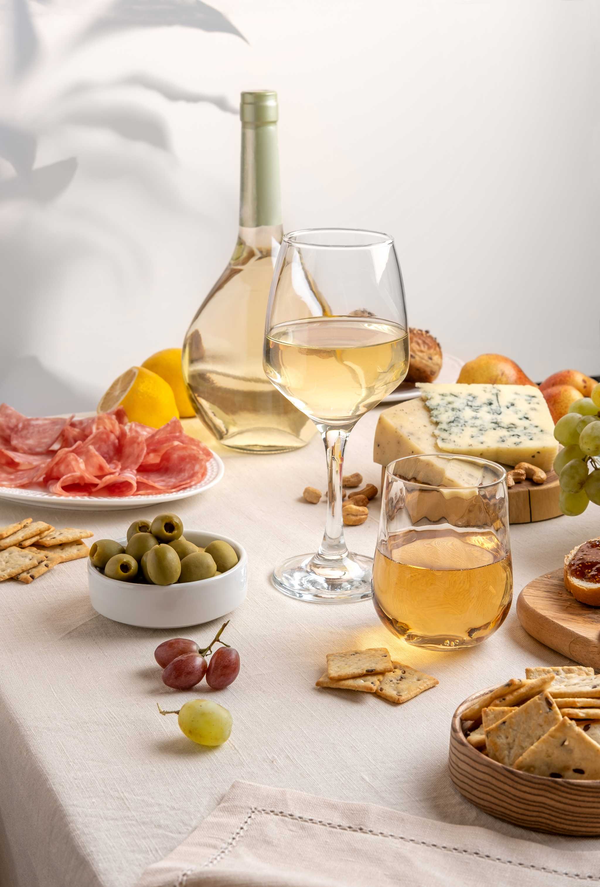 Two glasses and one bottle of wine plus assorted cheese, meats, crackers, and olives on a white tablecloth.