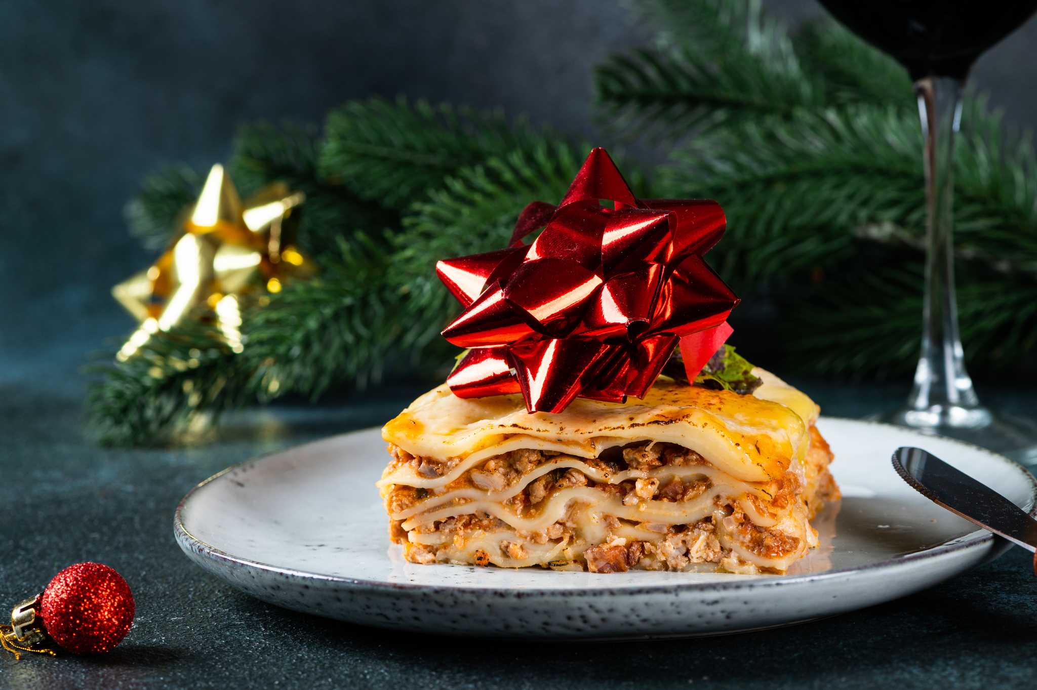 Plate of lasagna with a red gift bow on top and pine bough in the background.