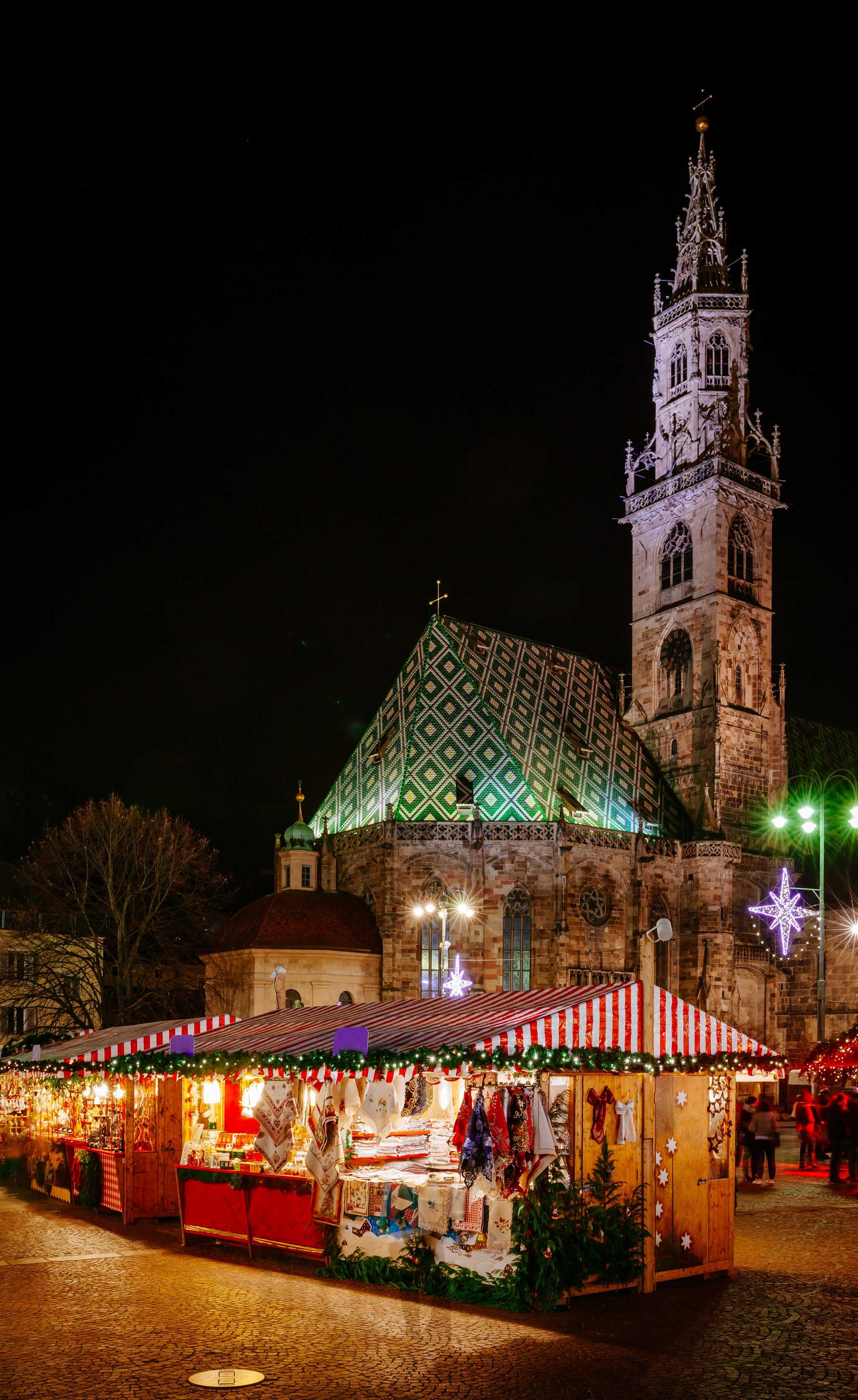 Nighttime shot of a warmly lit Christmas market stall in front of the cathedral in Bolzano.