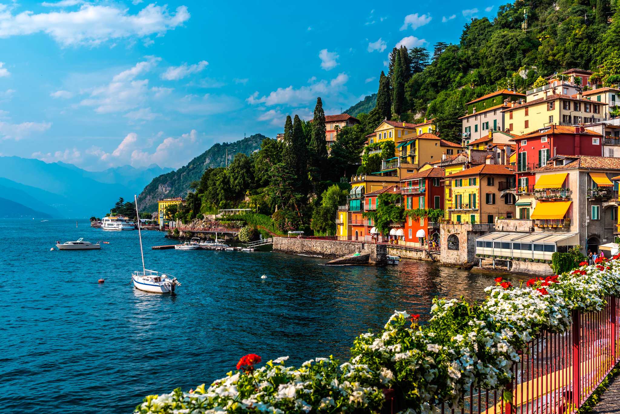 Flowers line a promenade on Lake Como that hugs the shore with colorful buildings.