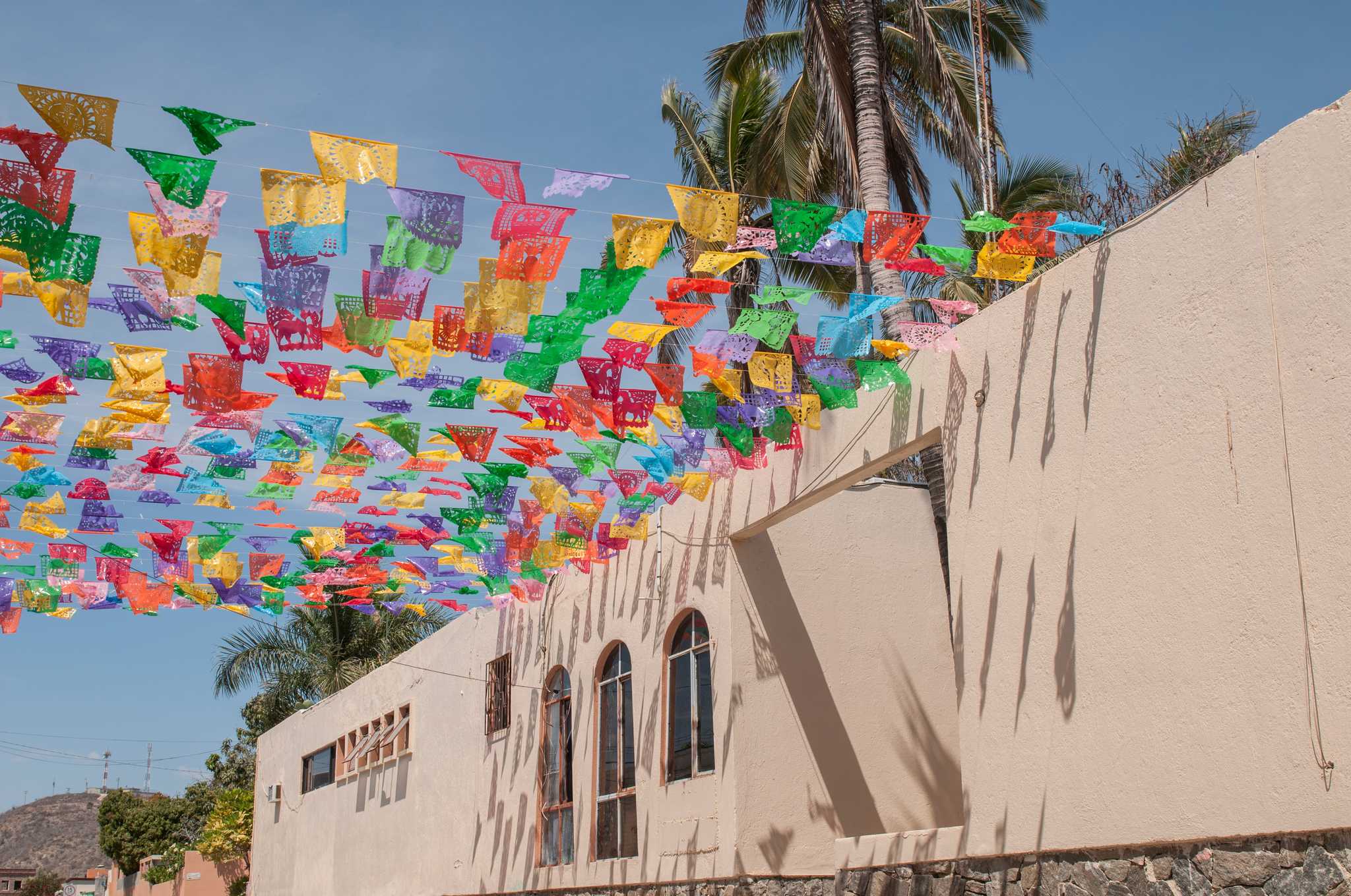 Colorful flags over a street in Todos Santos in front of a beige building.