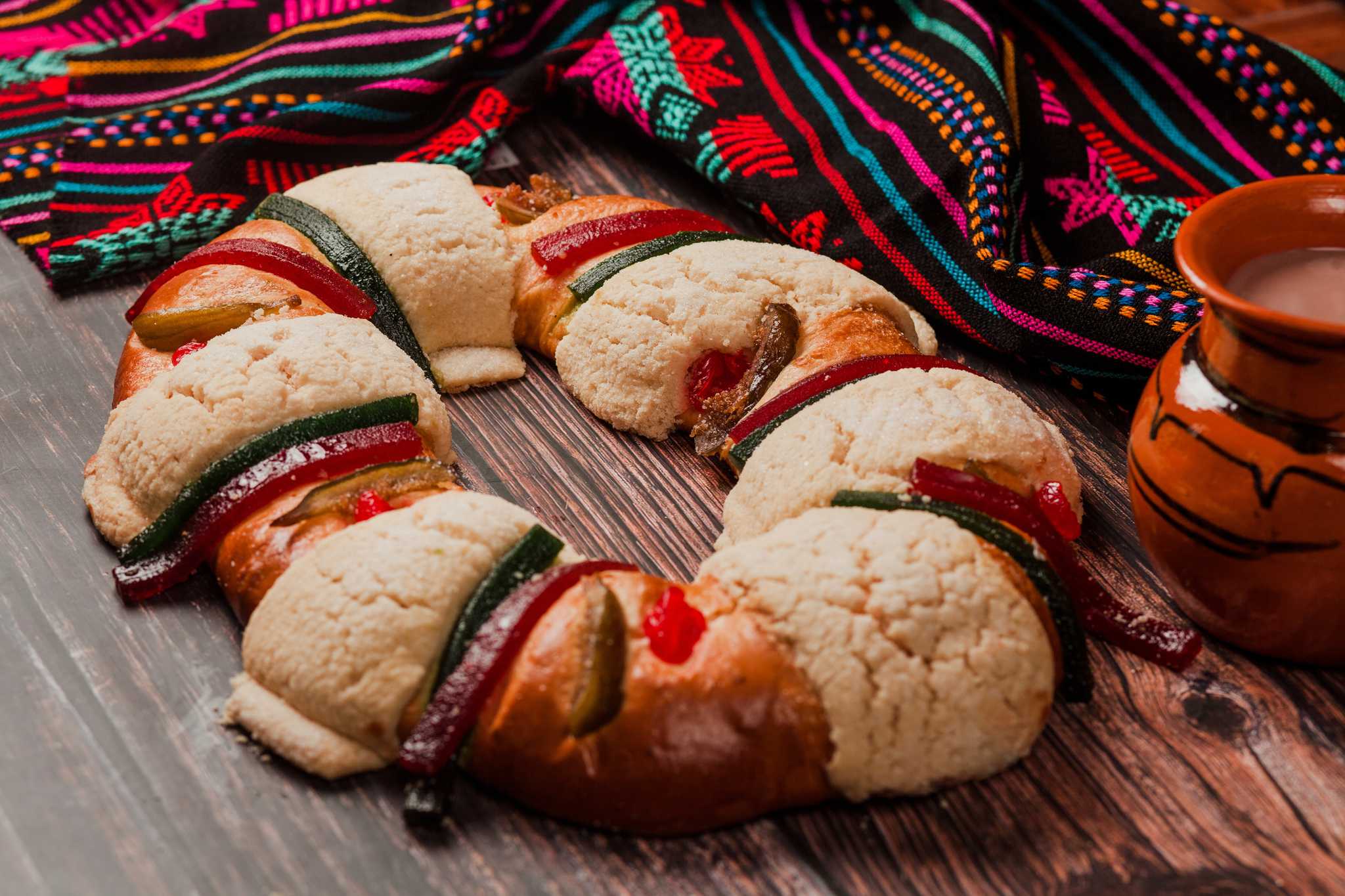Rosca de reyes topped with candied fruit beside a clay mug and woven Mexican textile.