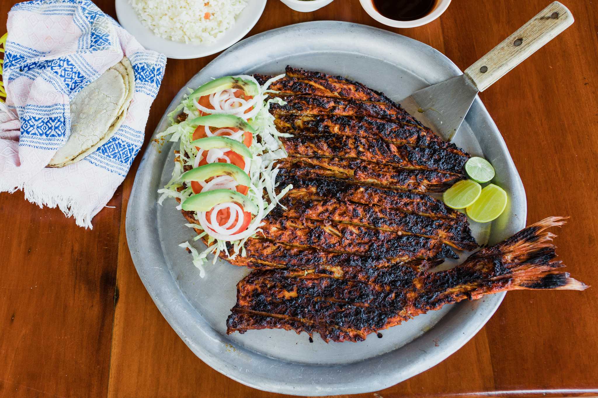 Plate of charred grilled fish, with lime, avocado, and tomatoes on the side.