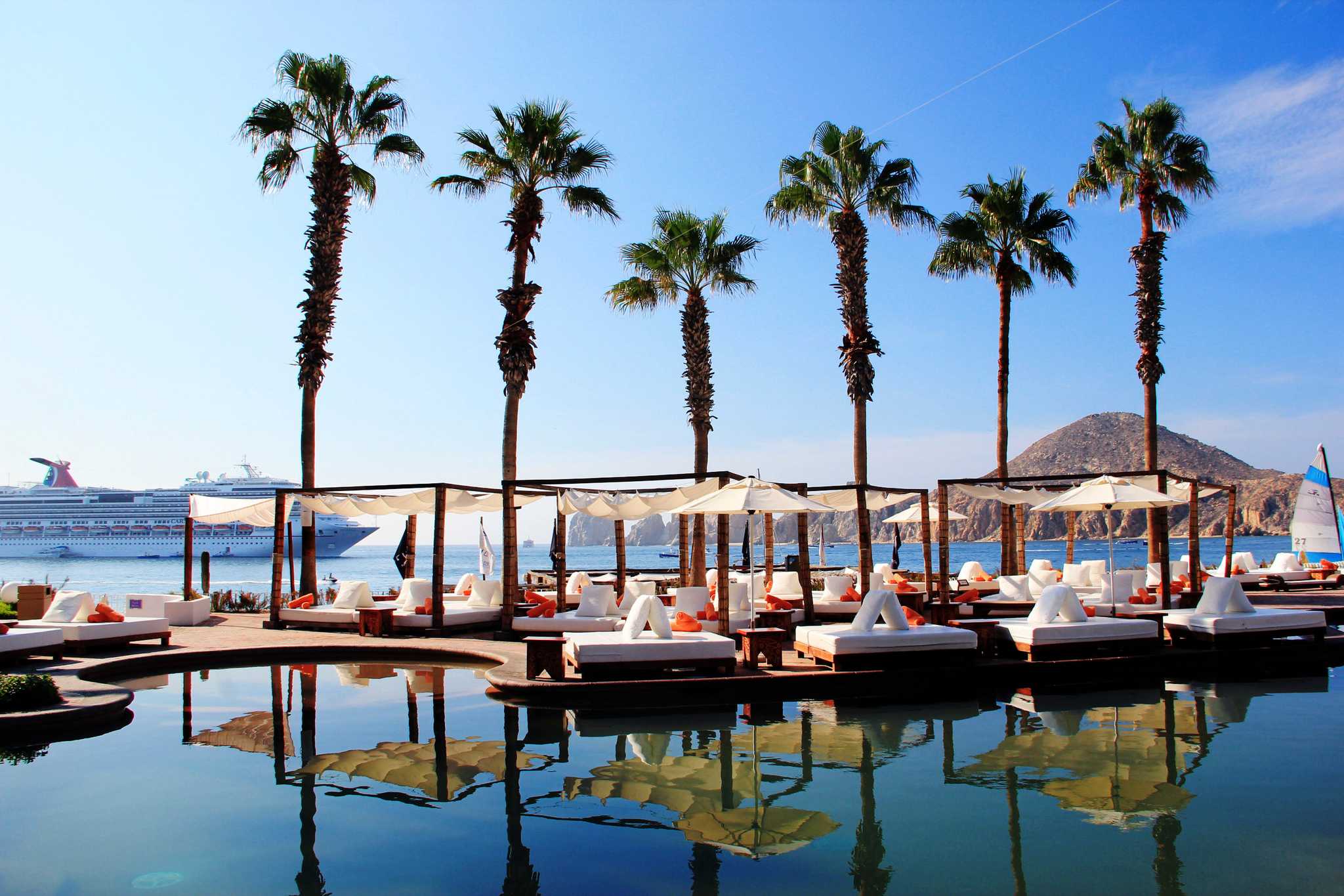 Lounge chairs and palm trees on the water in Los Cabos, a cruise ship in the background.