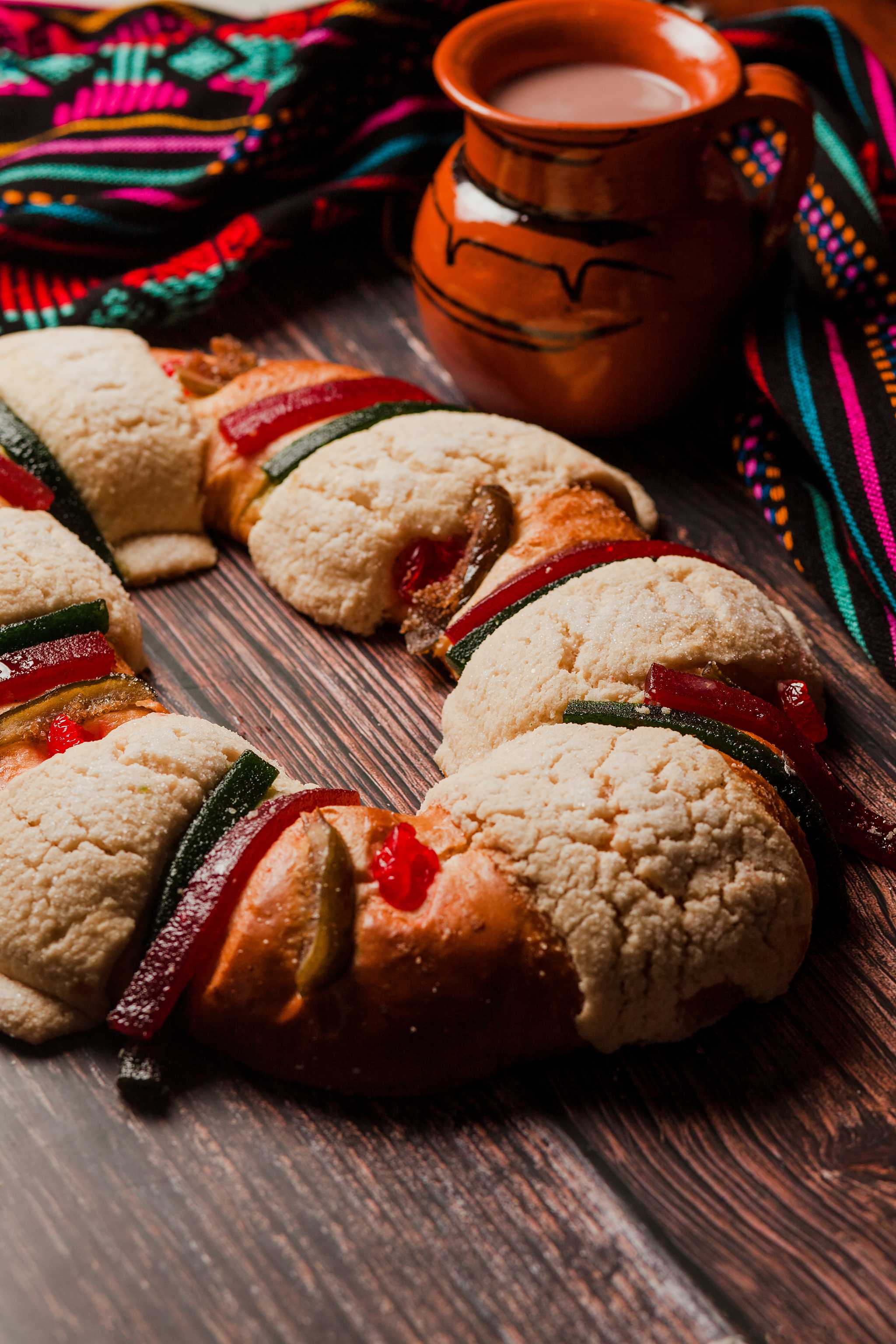 Rosca de reyes cake beside a clay mug of hot chocolate and colorful fabric.