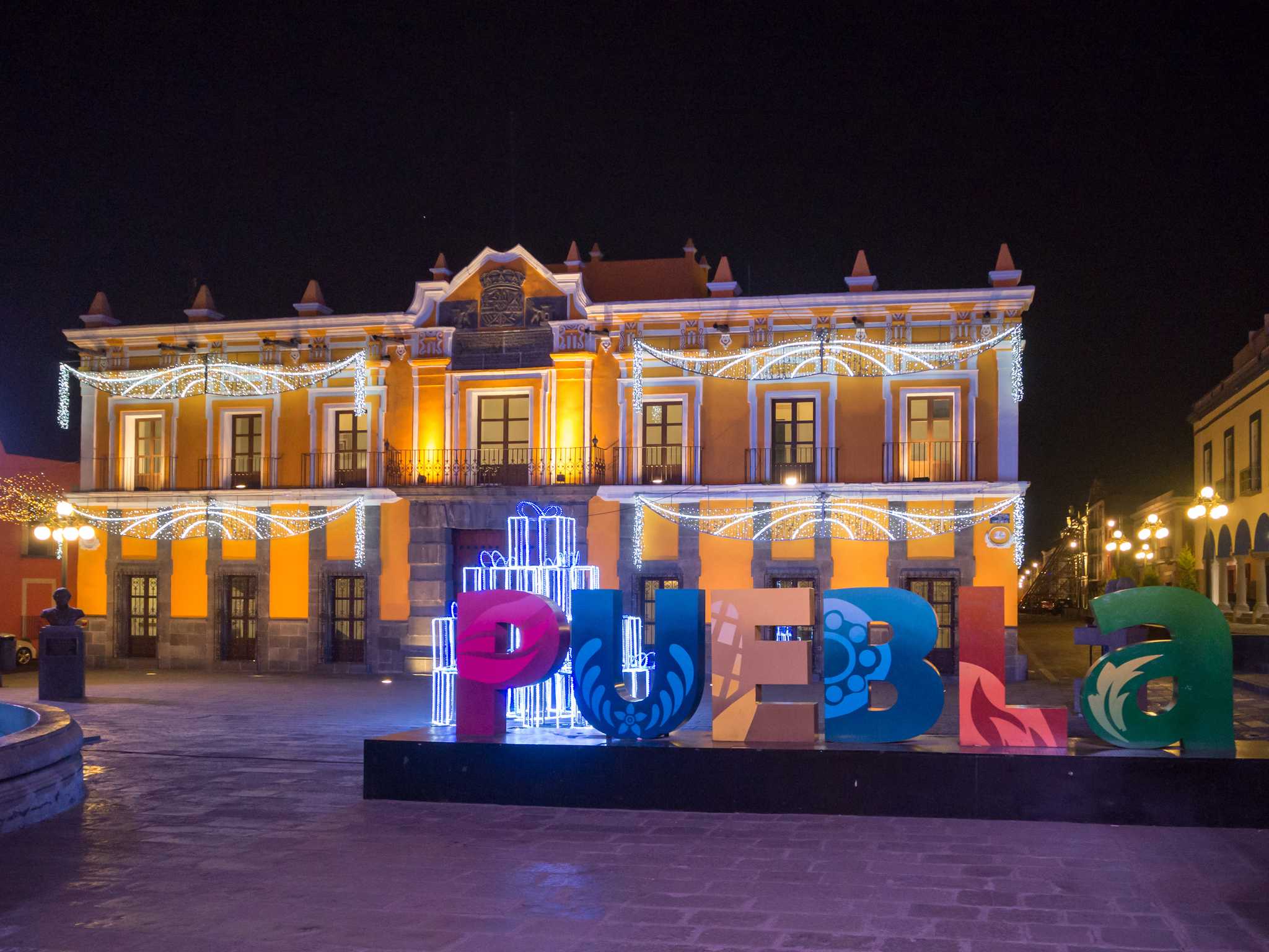 Colorful Puebla City sign in front of a yellow building decorated with Christmas lights.