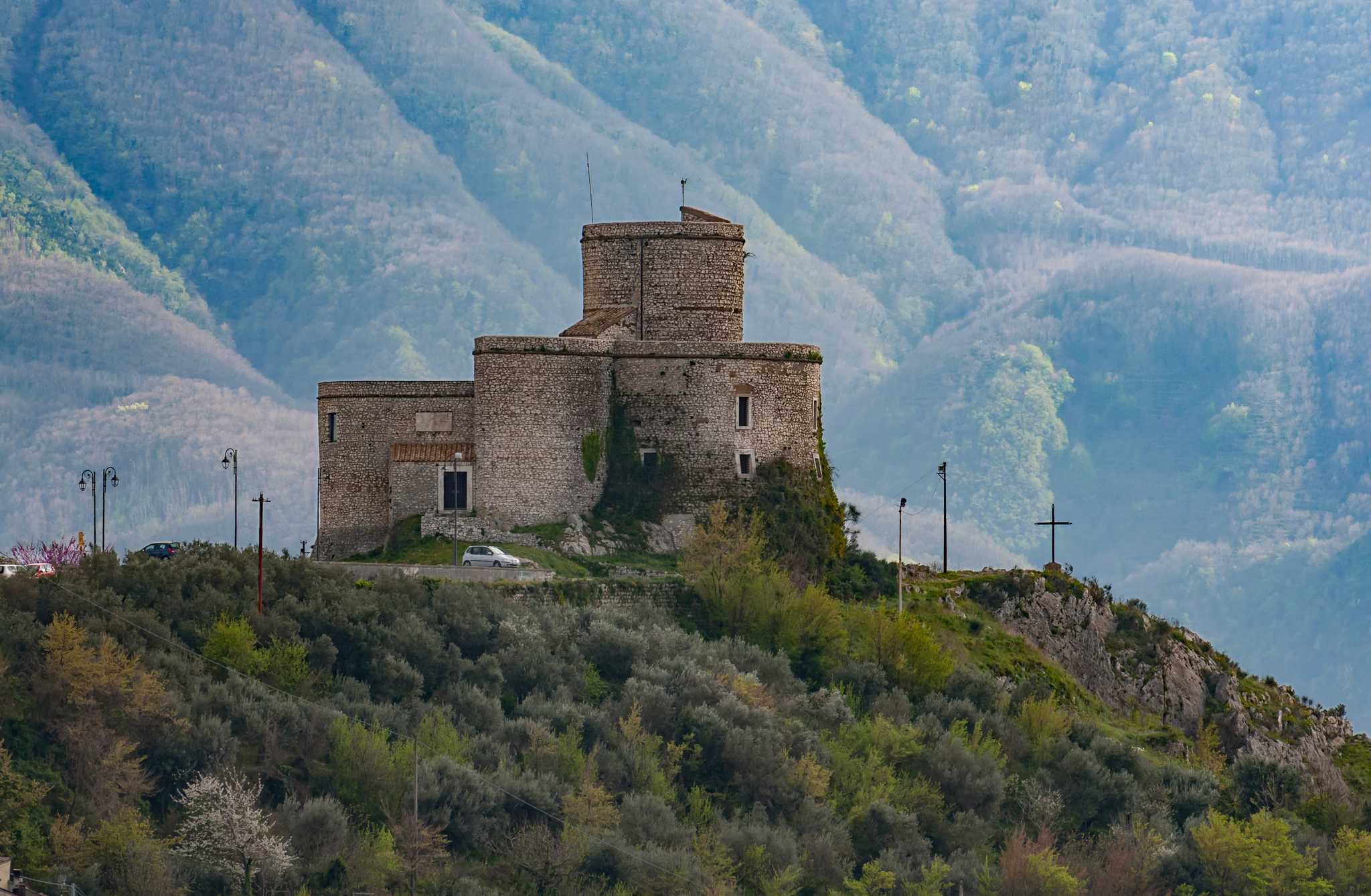 Stone castle with a round tower on a wooded ridge before hazy mountains.