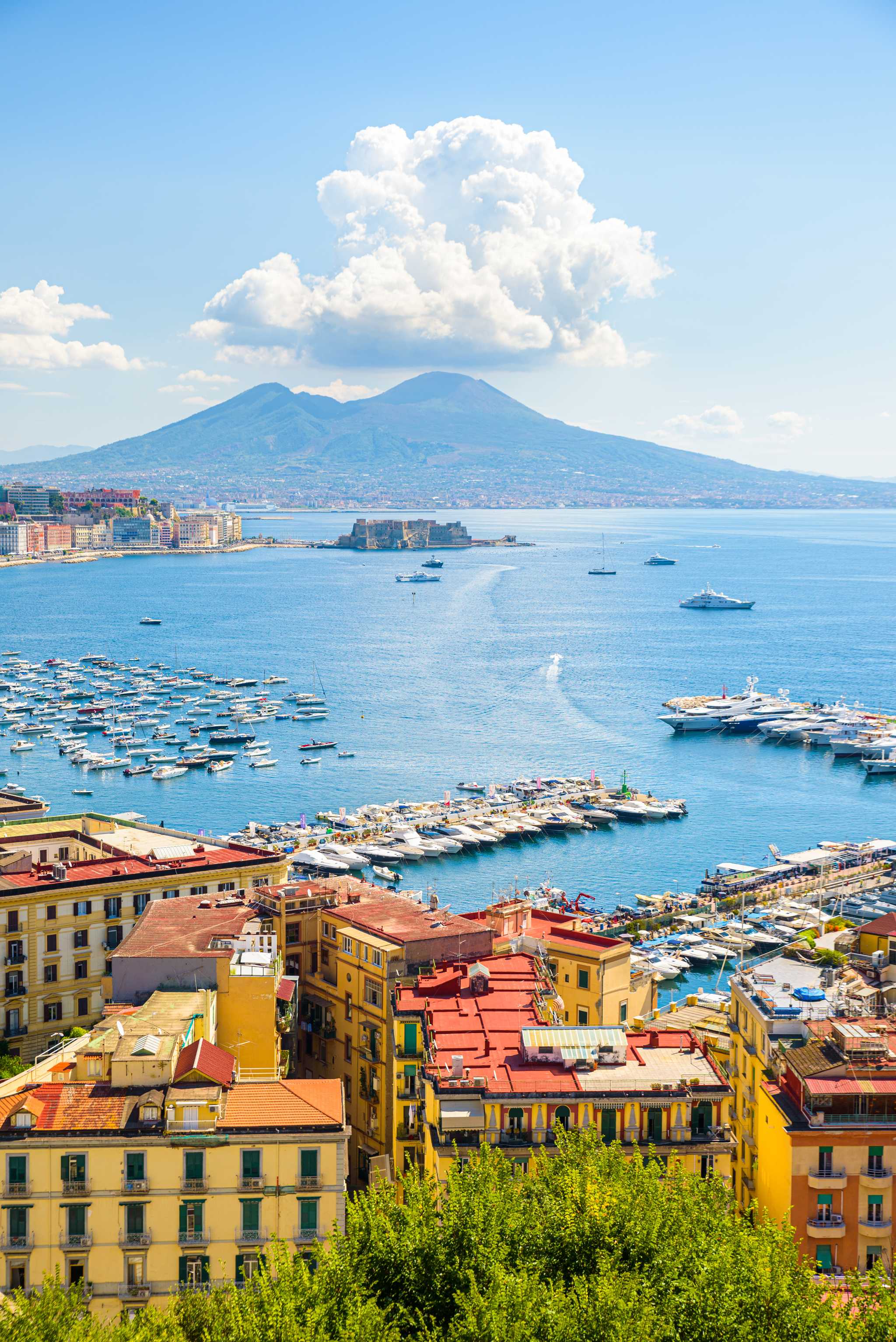Boats fill the Bay of Naples below Mount Vesuvius, with the city's rooftops in the foreground.