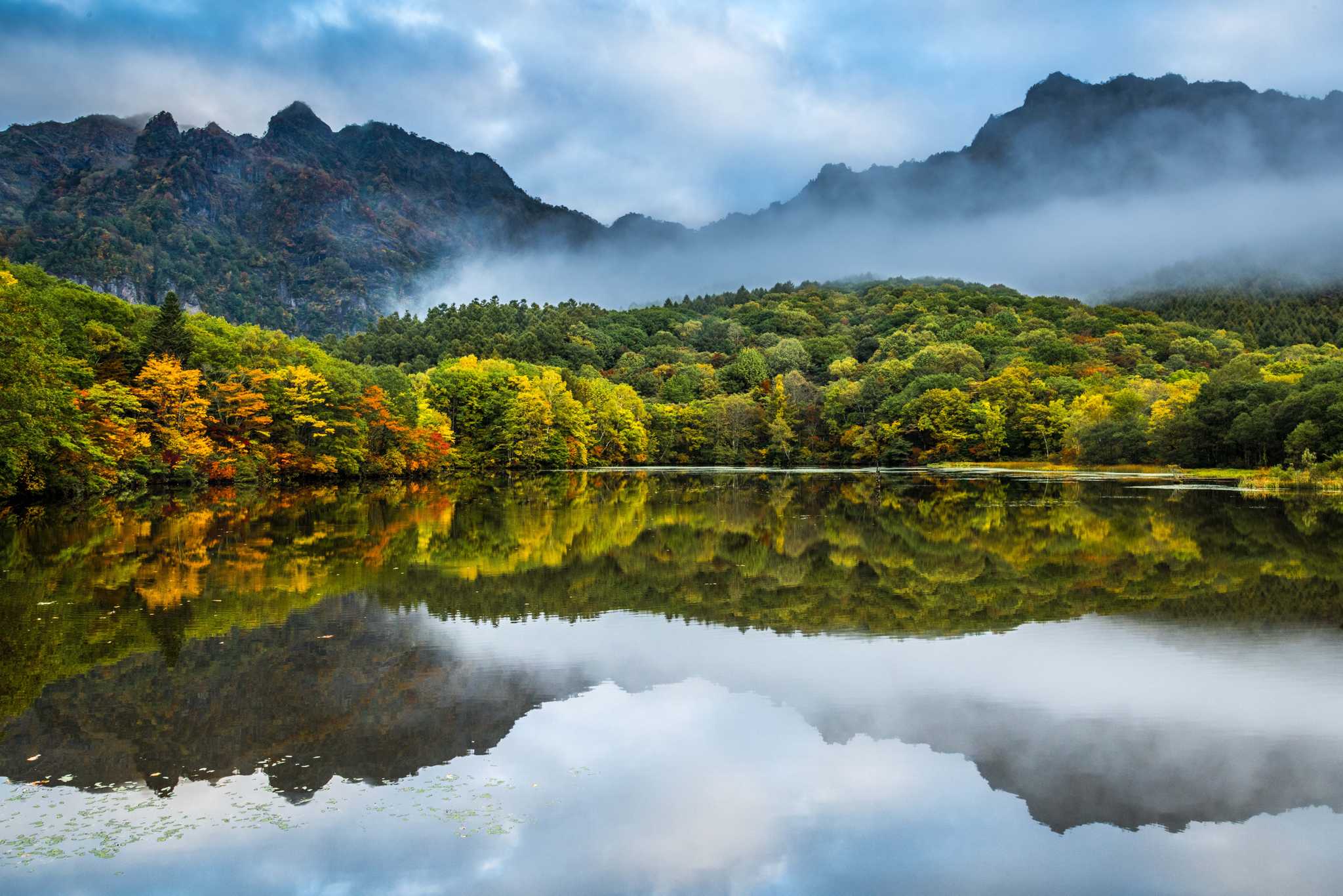 Misty forested mountains reflected in a still lake with autumn foliage at the shore.