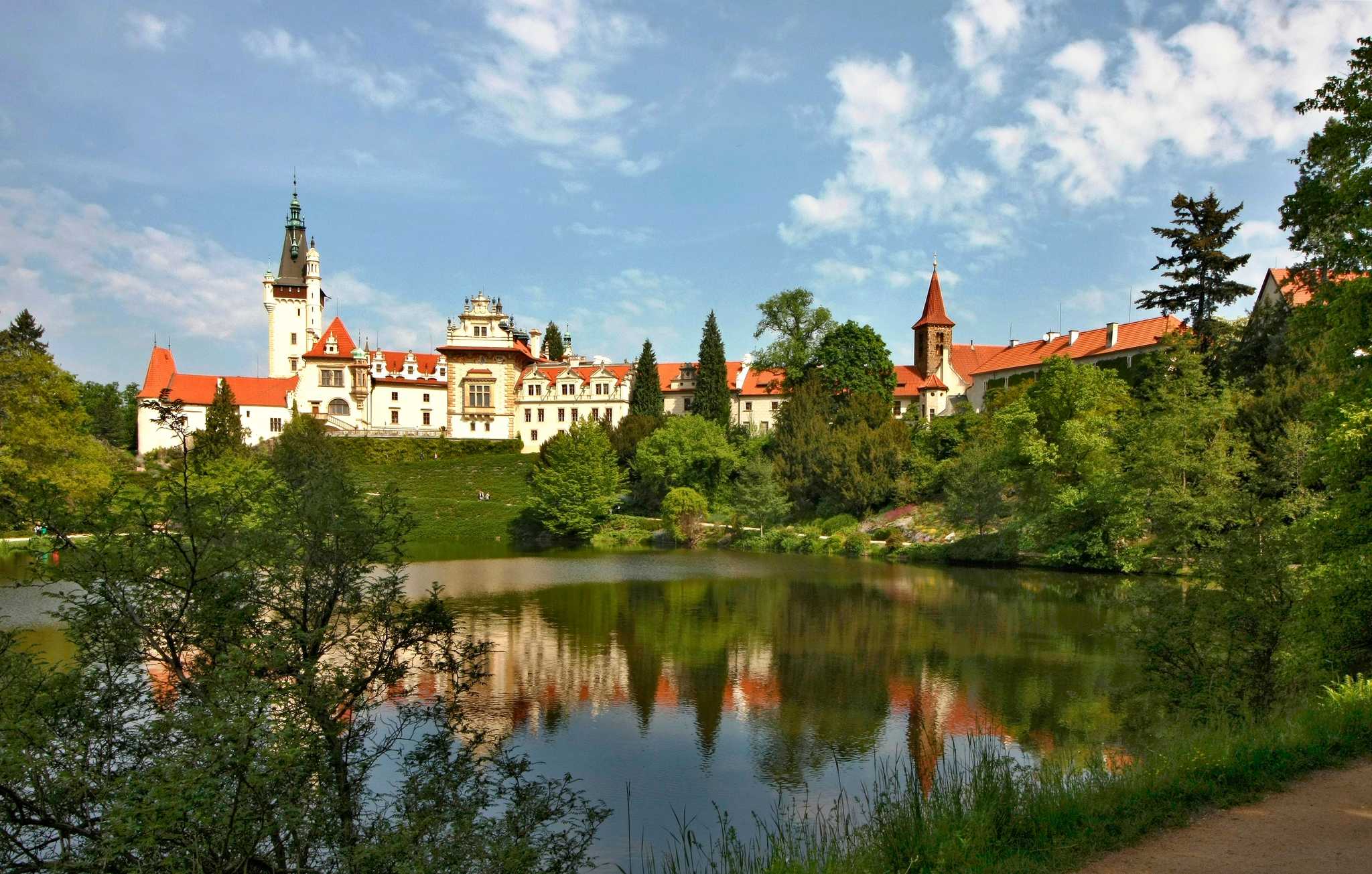 A Baroque-style castle complex spans the shores of a small lake surrounded by greenery.