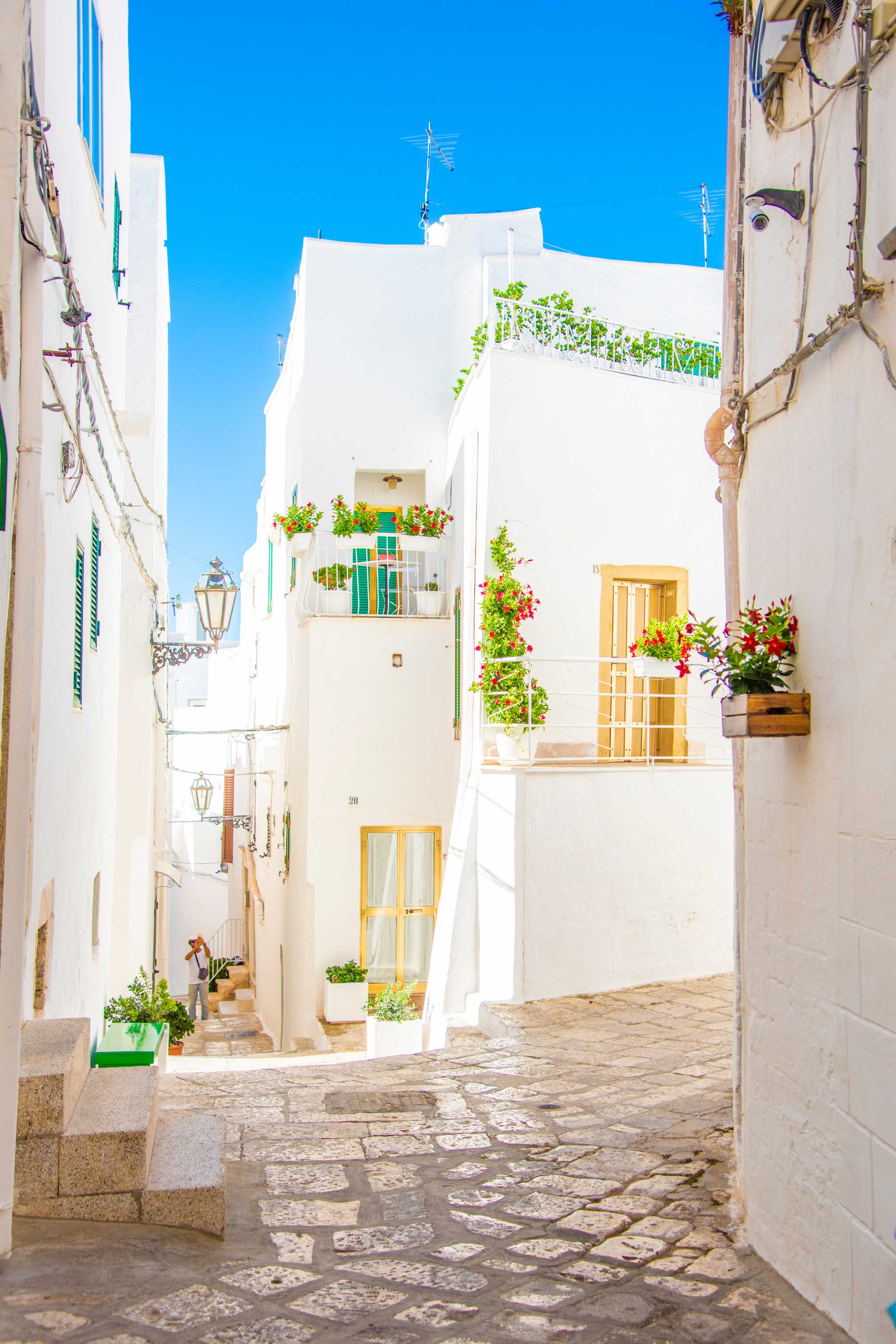 Cobblestone alleyway in Ostuni surrounded by bright white buildings with plants under a blue sky.