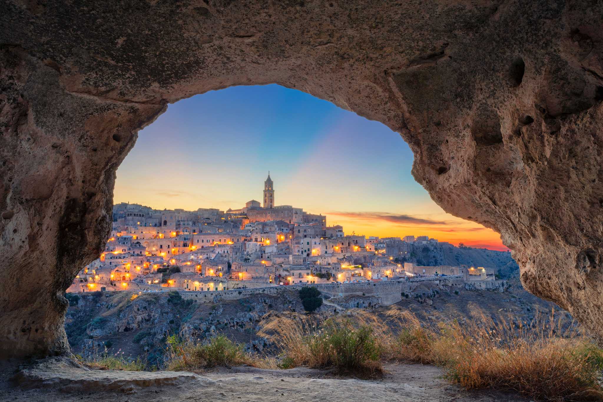 A cave mouth framing Matera at sunset, with warm lights dotting the city’s windows.