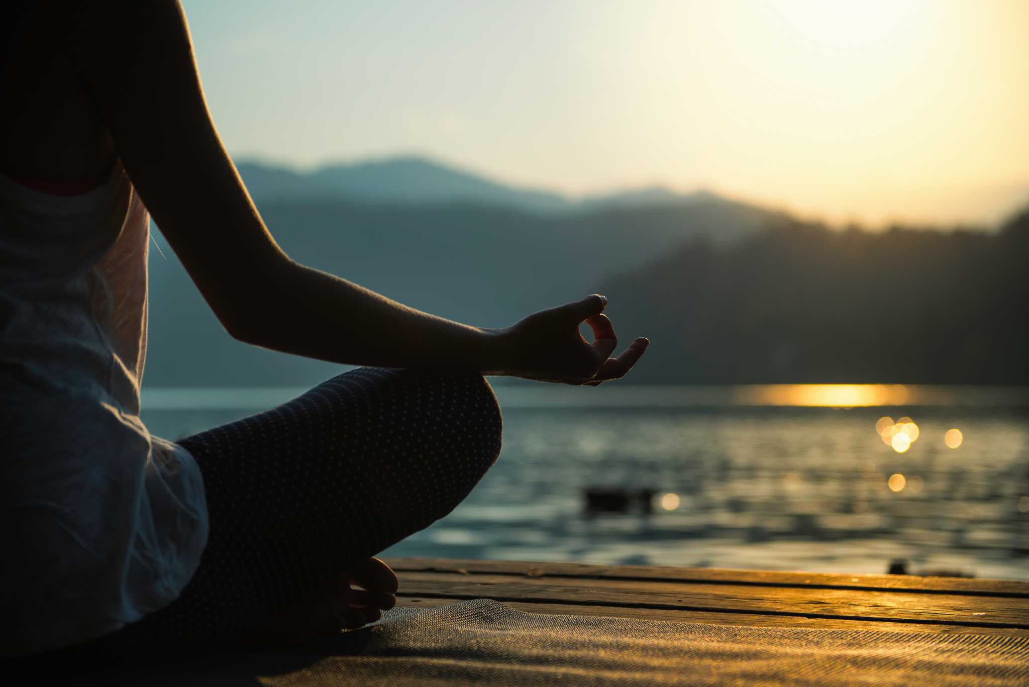 A person meditating cross-legged on a wooden dock beside a calm lake.