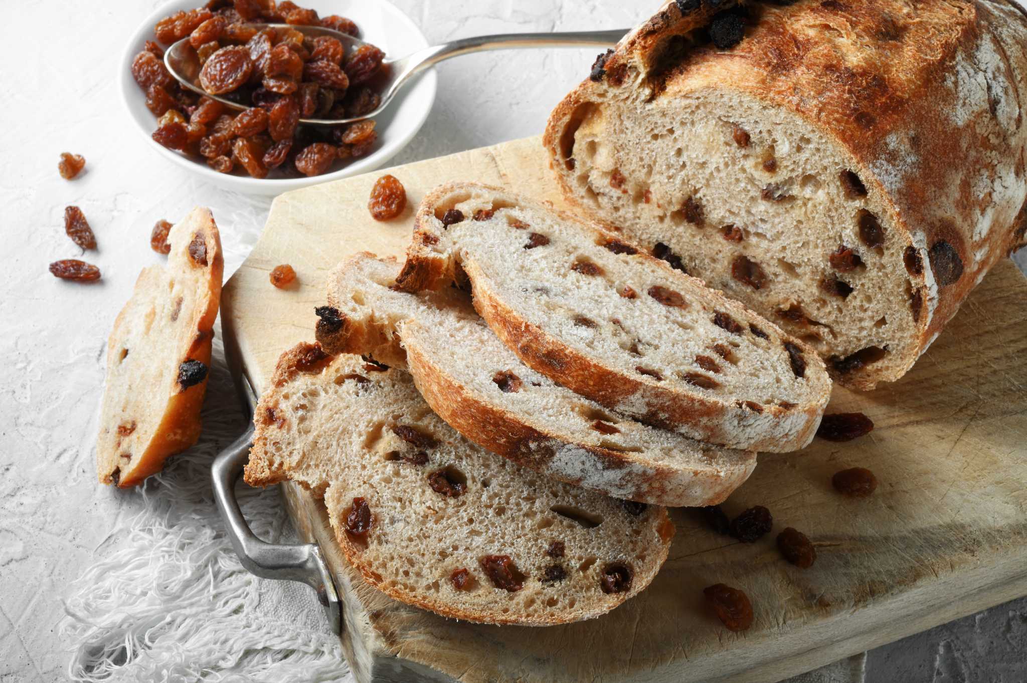 A sliced loaf of raisin bread on a wooden board, raisins scattered around.