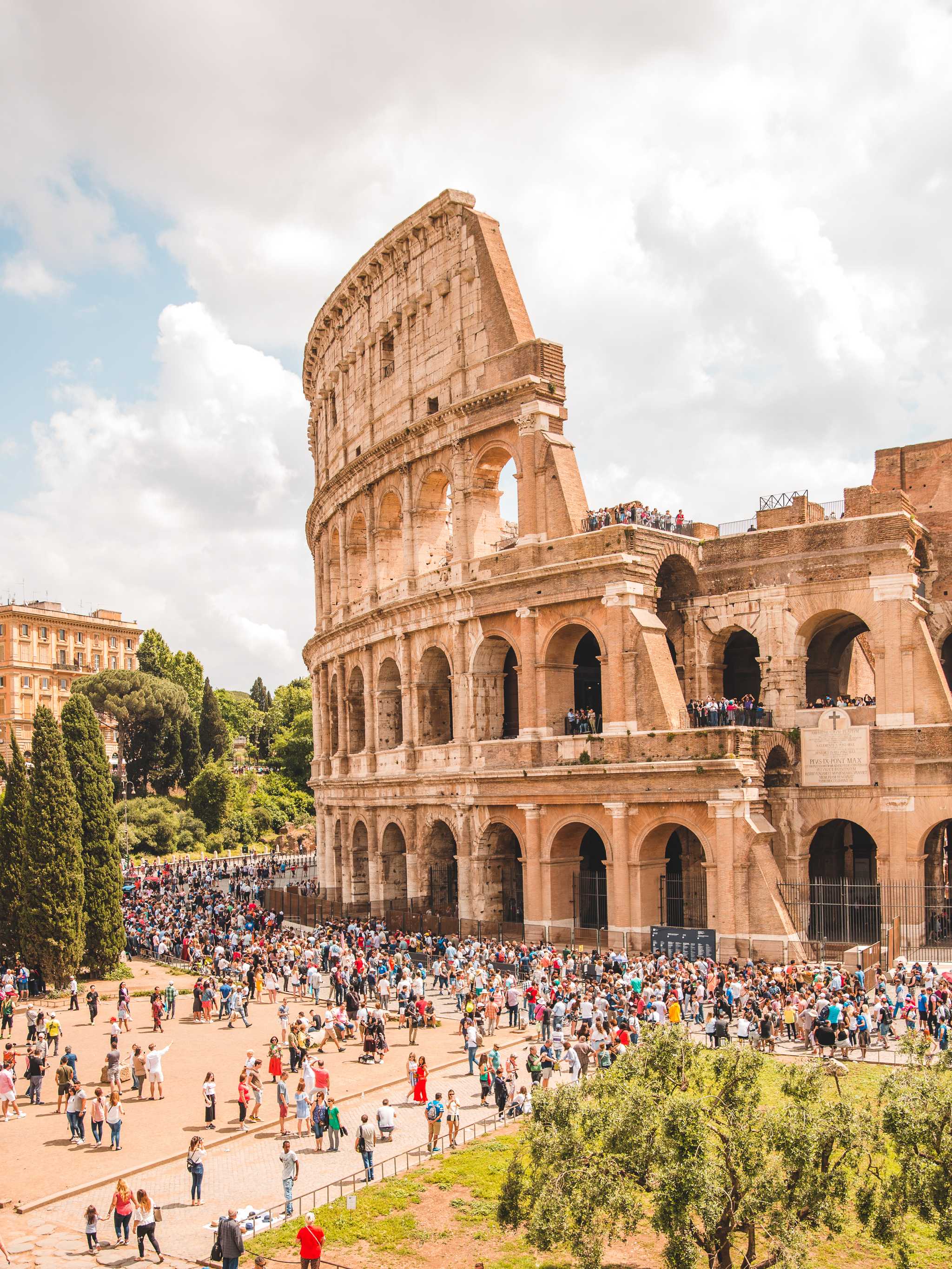 The Colosseum in Rome, its tiered stone arches rising above crowds of visitors gathered below.
