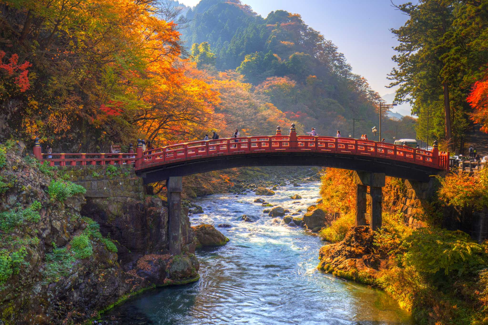 A red bridge spans a small river with a forested mountain in the background.