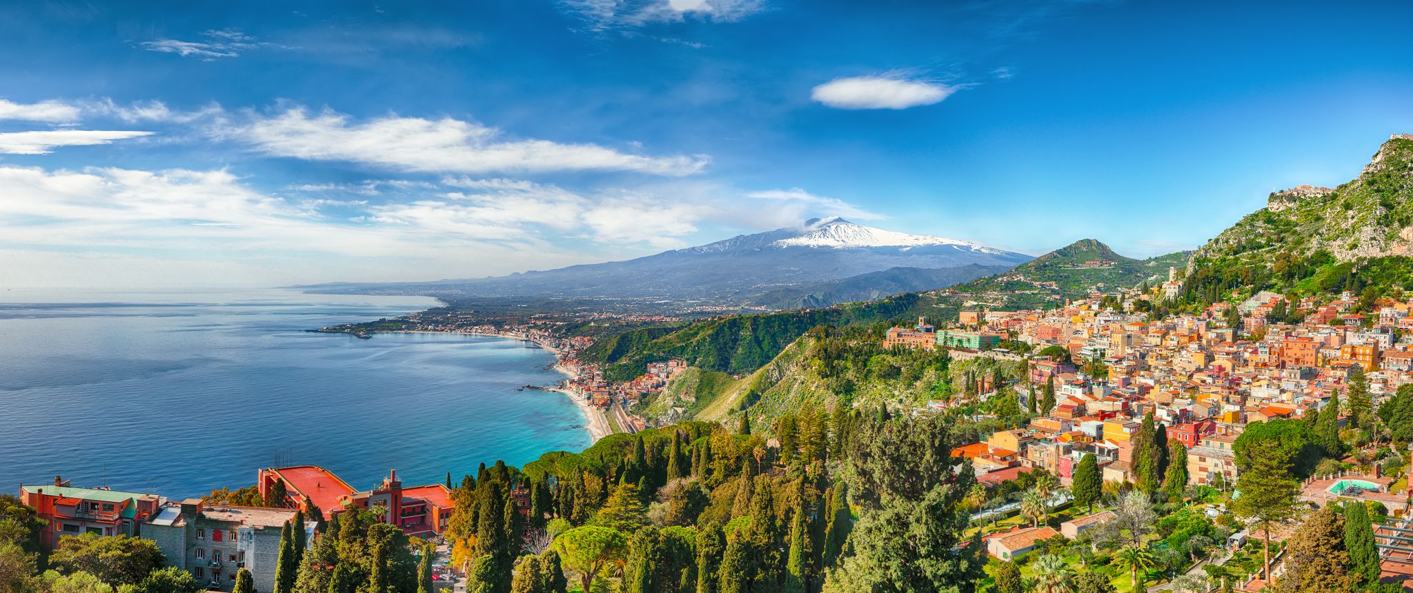 Taormina's colorful hillside town overlooking a turquoise bay with snow-capped Mount Etna beyond.