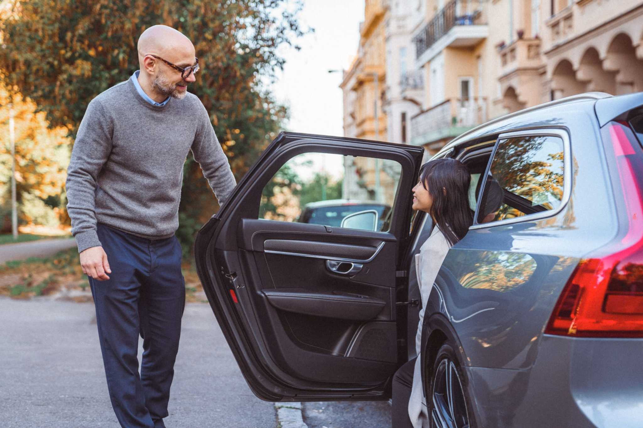 A driver holding the car door open for a smiling passenger on a tree-lined city street