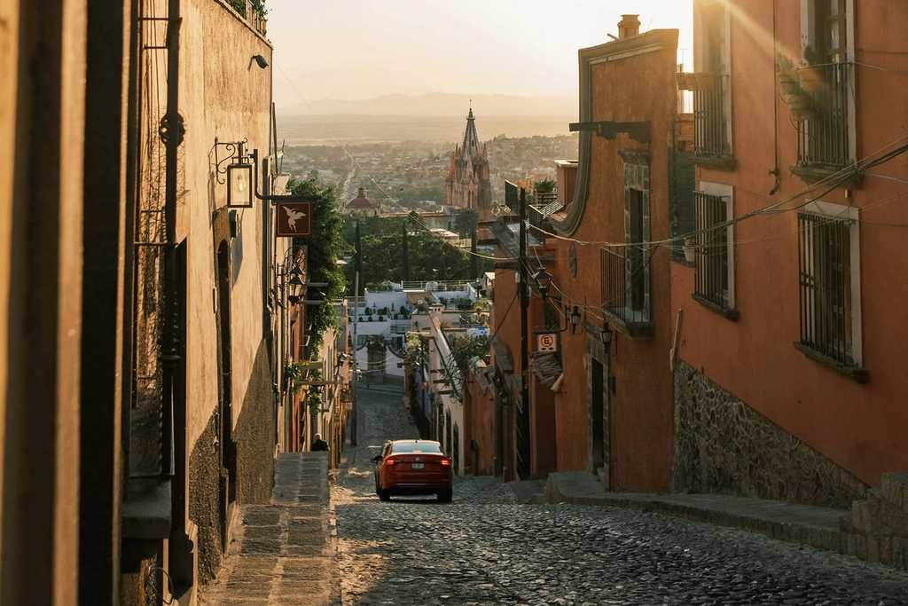 A car on a cobblestone street in San Miguel de Allende at sunset, with the city's pink church in the distance