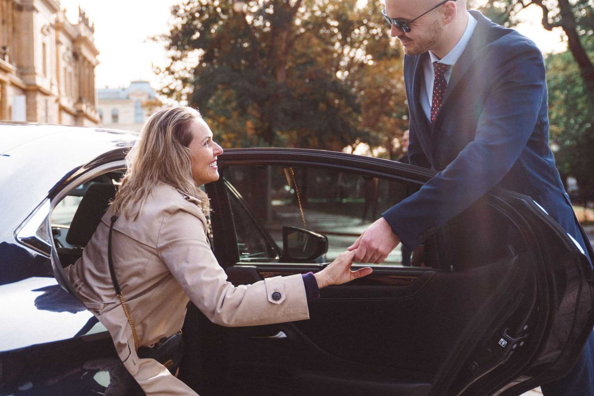 A suited chauffeur holding the door and offering his hand to a smiling passenger