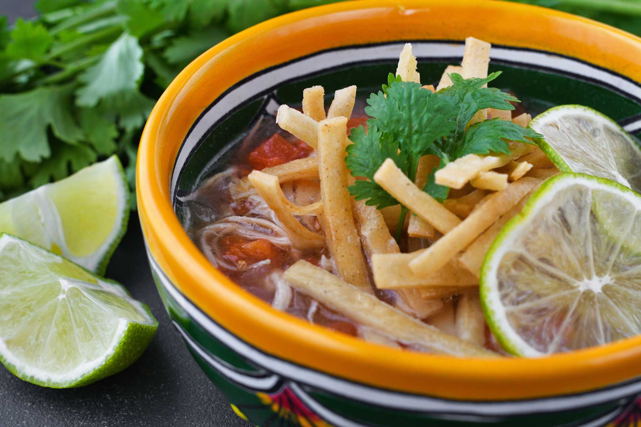 A bowl of sopa de lima with coriander and fried tortilla strips served with a lime wedge.