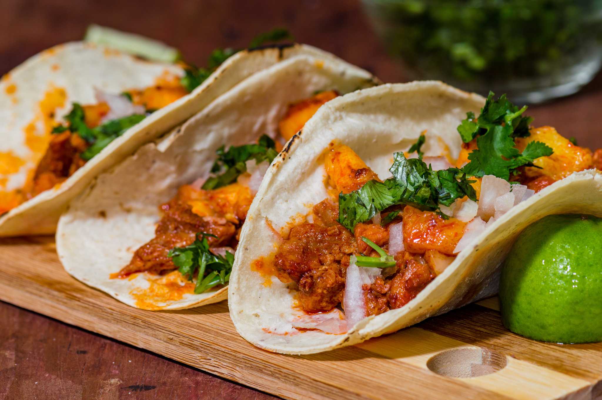 Three tacos al pastor topped with cilantro next to a wedge of lime on a cutting board.