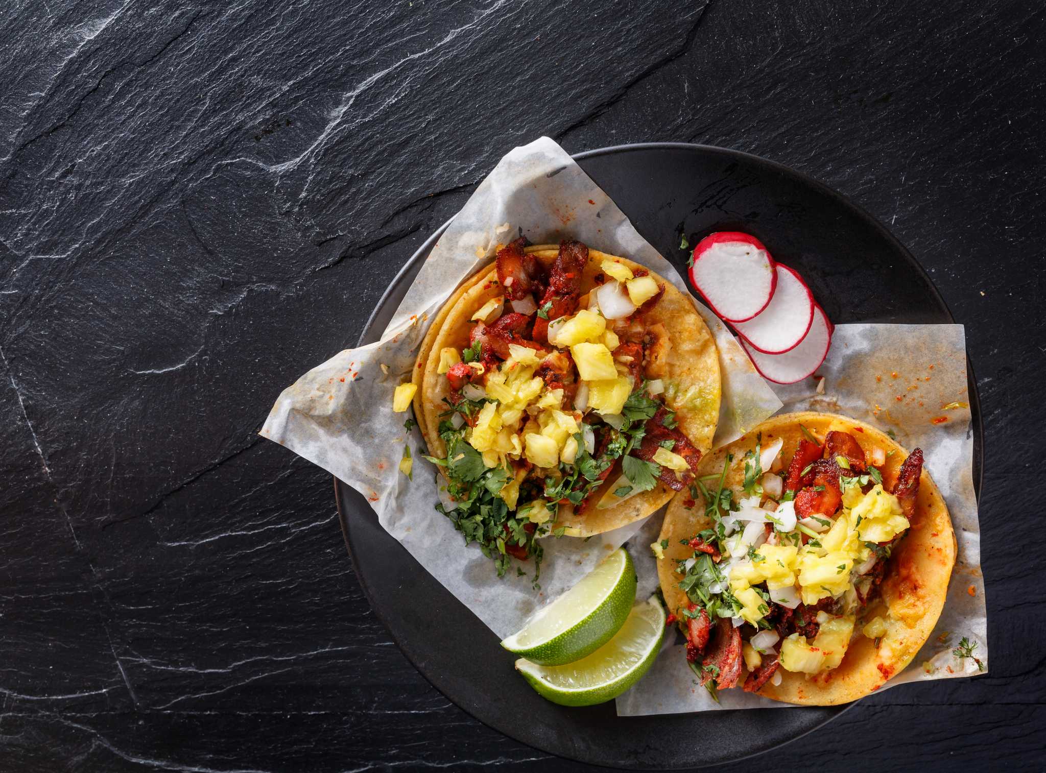 Two tacos al pastor on a black plate and served with lime and radishes.