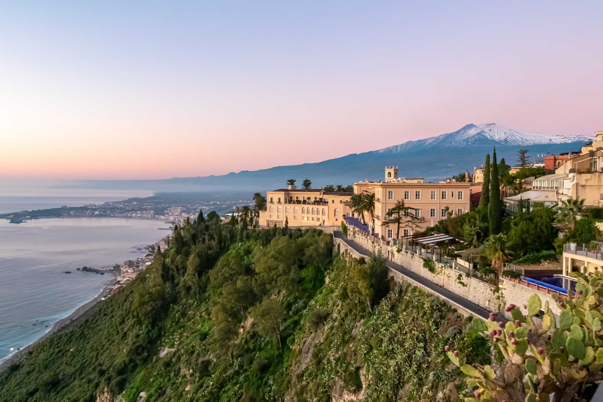 Taormina's clifftop luxury hotel San Domenico Palace above the sea at dusk, snow-capped Mount Etna beyond.