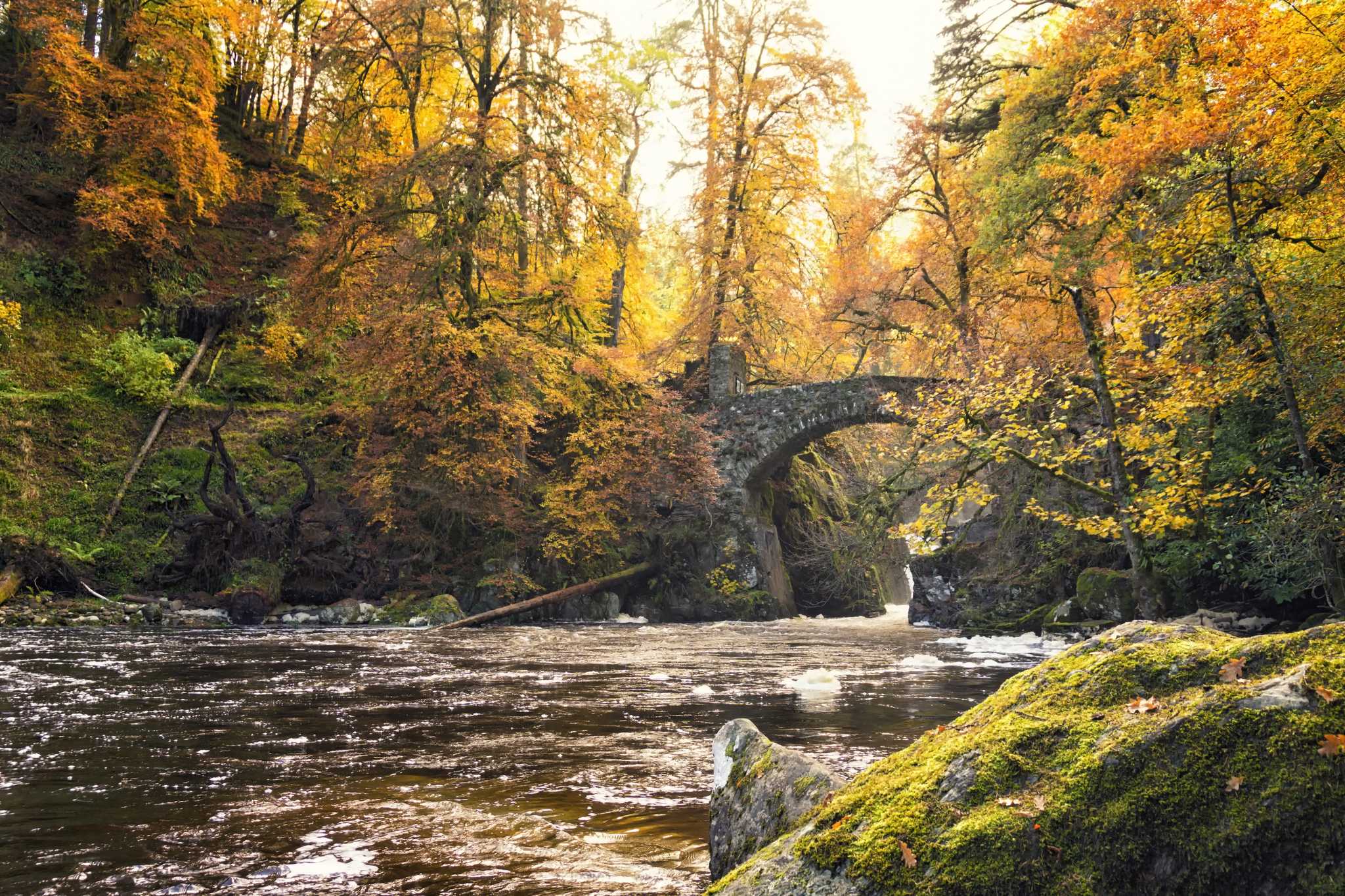 An ancient stone bridge spans a narrow riverbed with rushing water and rocks.