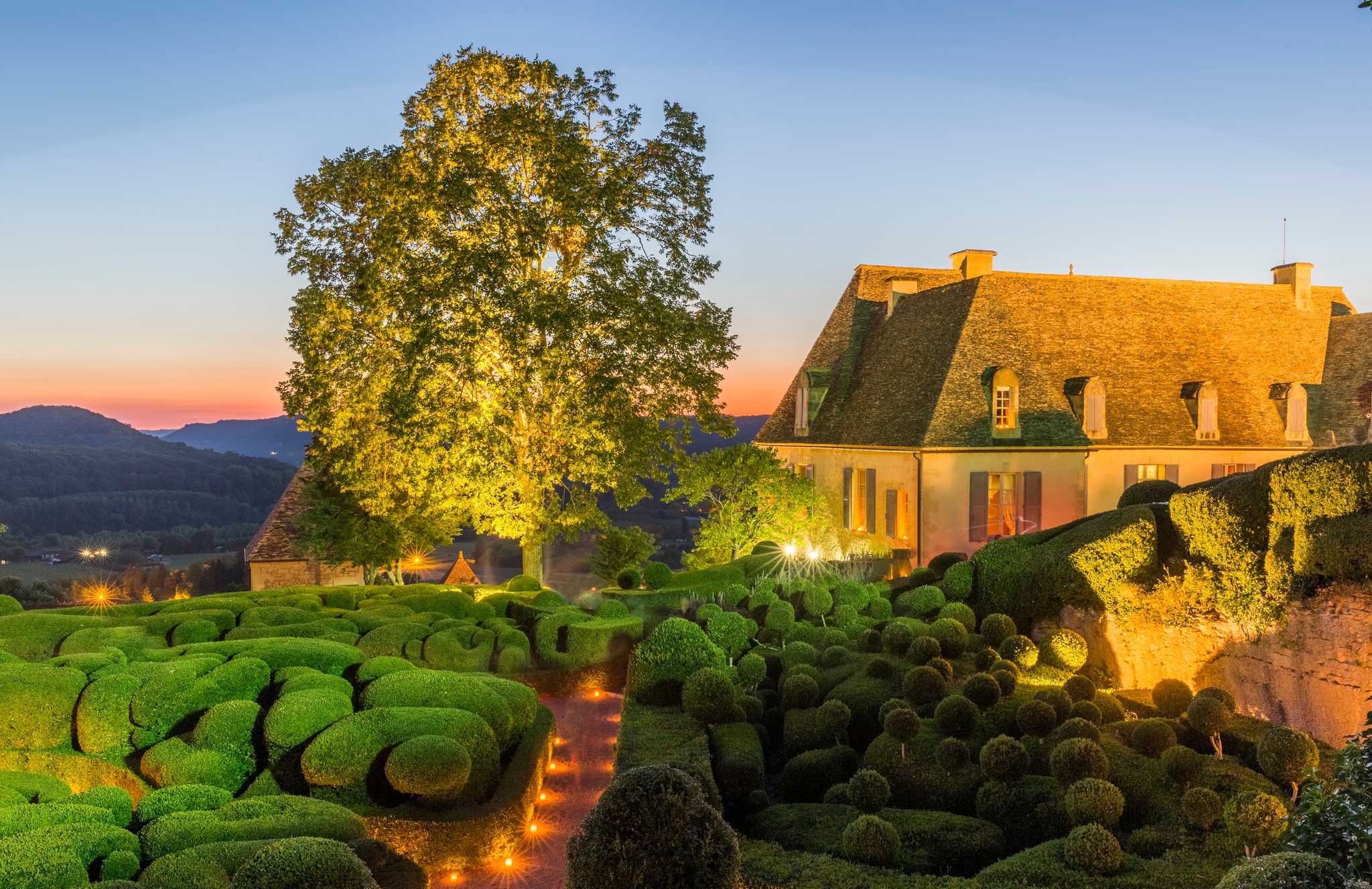 A garden of manicured bushes encircles a chateau with rolling hills in the background.