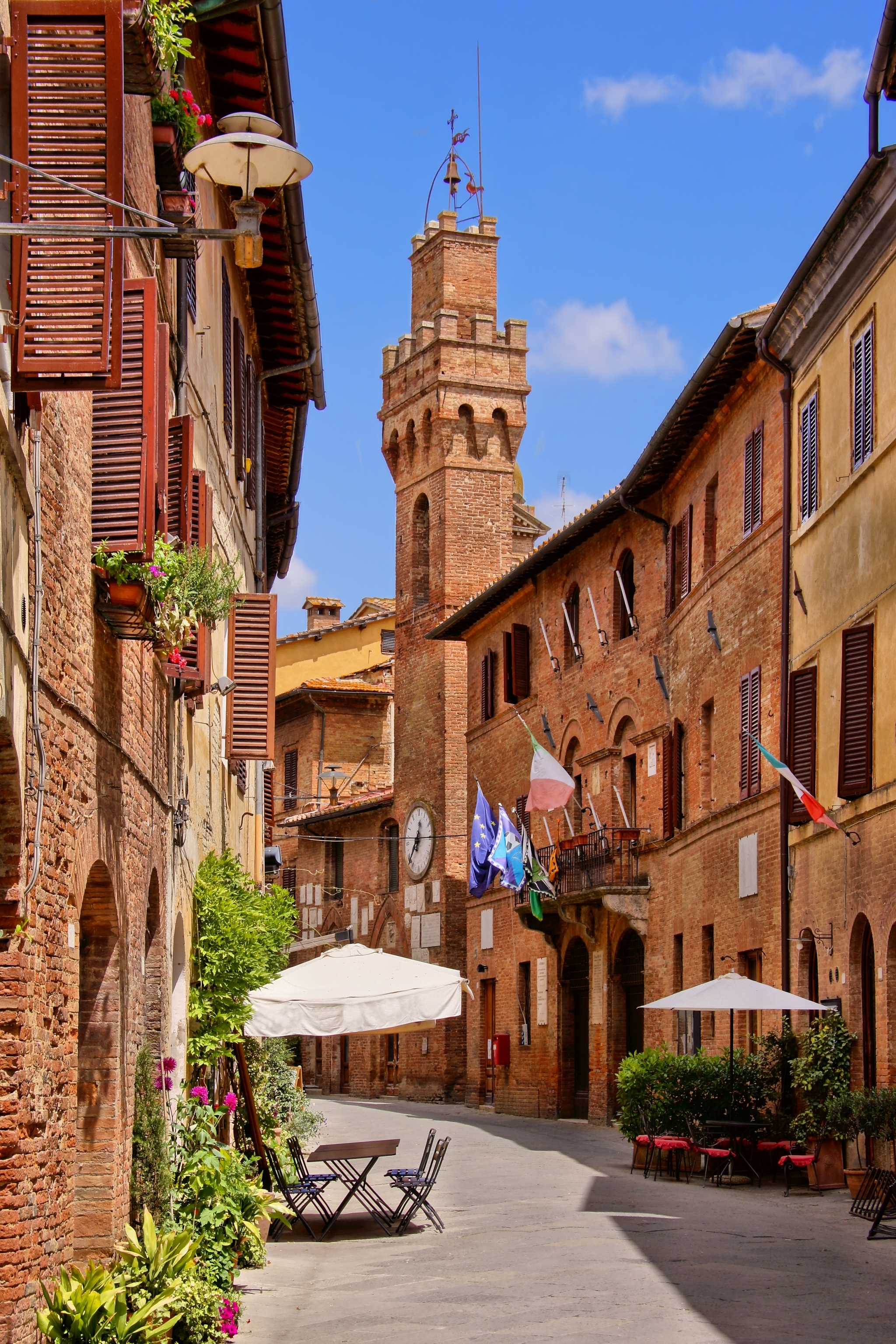 Siena's curved alleyway with red-stone buildings, one with a tall tower, tables and chairs in the walkway.