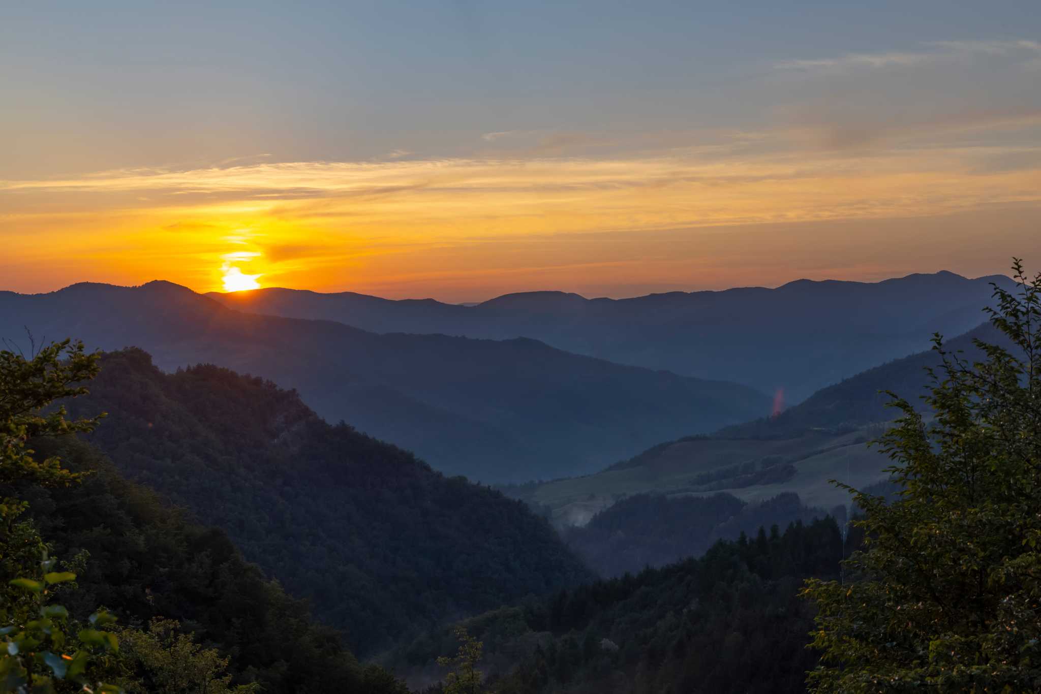 Rolling hills of Tuscany at dusk with a forest in the foreground and rows of mountains.