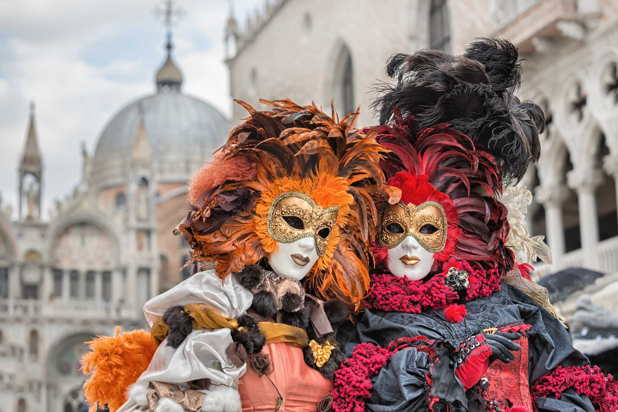 Two ornate feathered Venice Carnival masks and costumes with St Mark's Basilica in the background.